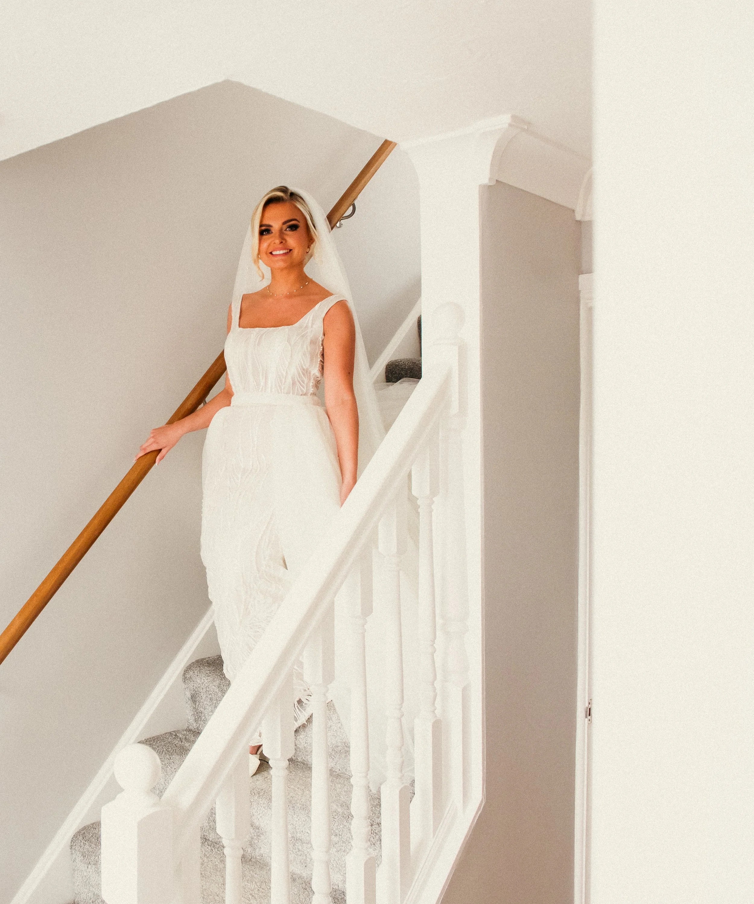 A bride in a white wedding dress standing on a staircase with a wooden handrail, smiling at the camera.