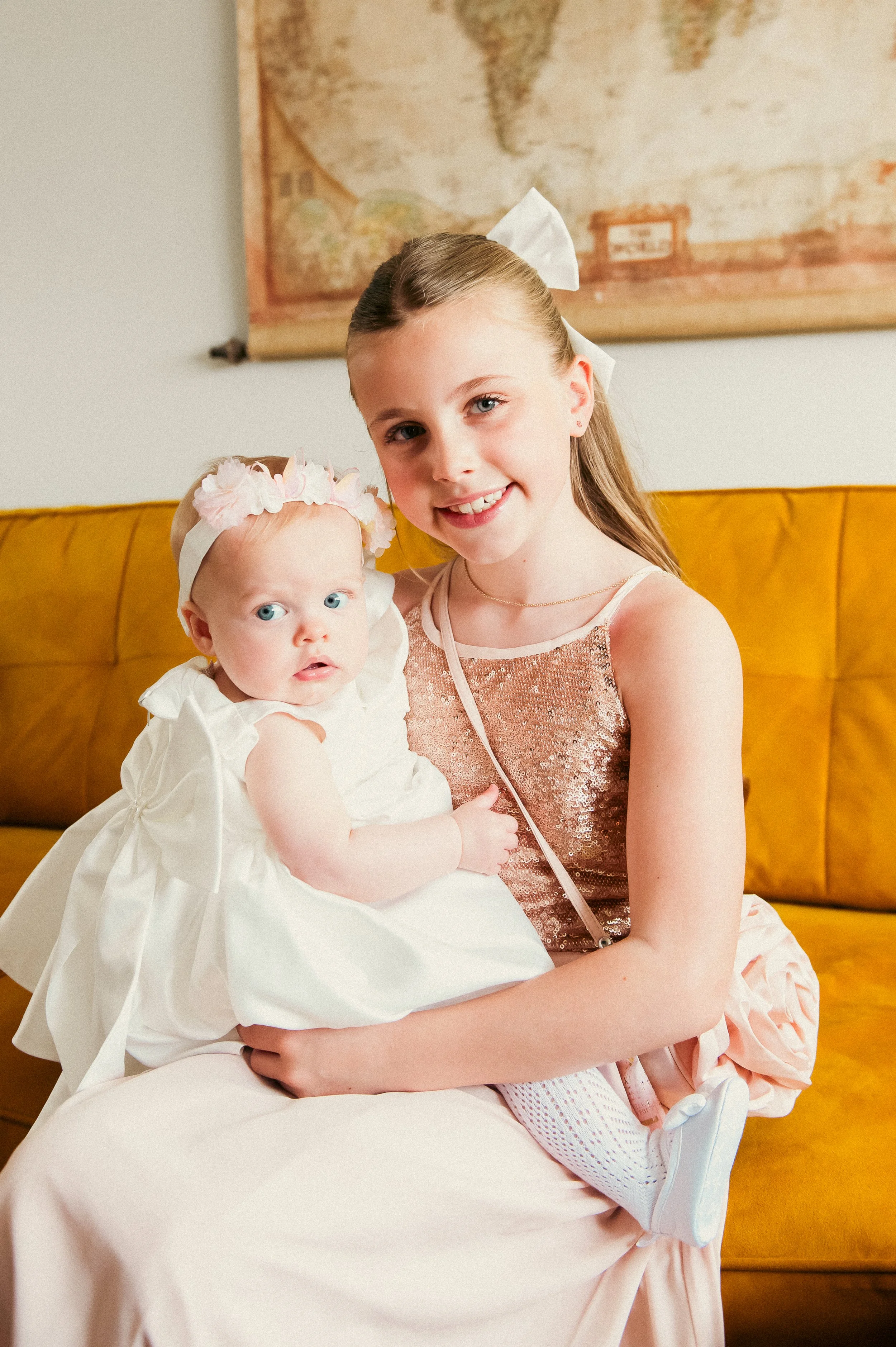 A young girl with long light brown hair, wearing a sleeveless dress with a sequined top and a bow in her hair, sitting on a mustard-yellow couch. She is holding a baby girl with blonde hair, blue eyes, wearing a white dress and a pink flower headband