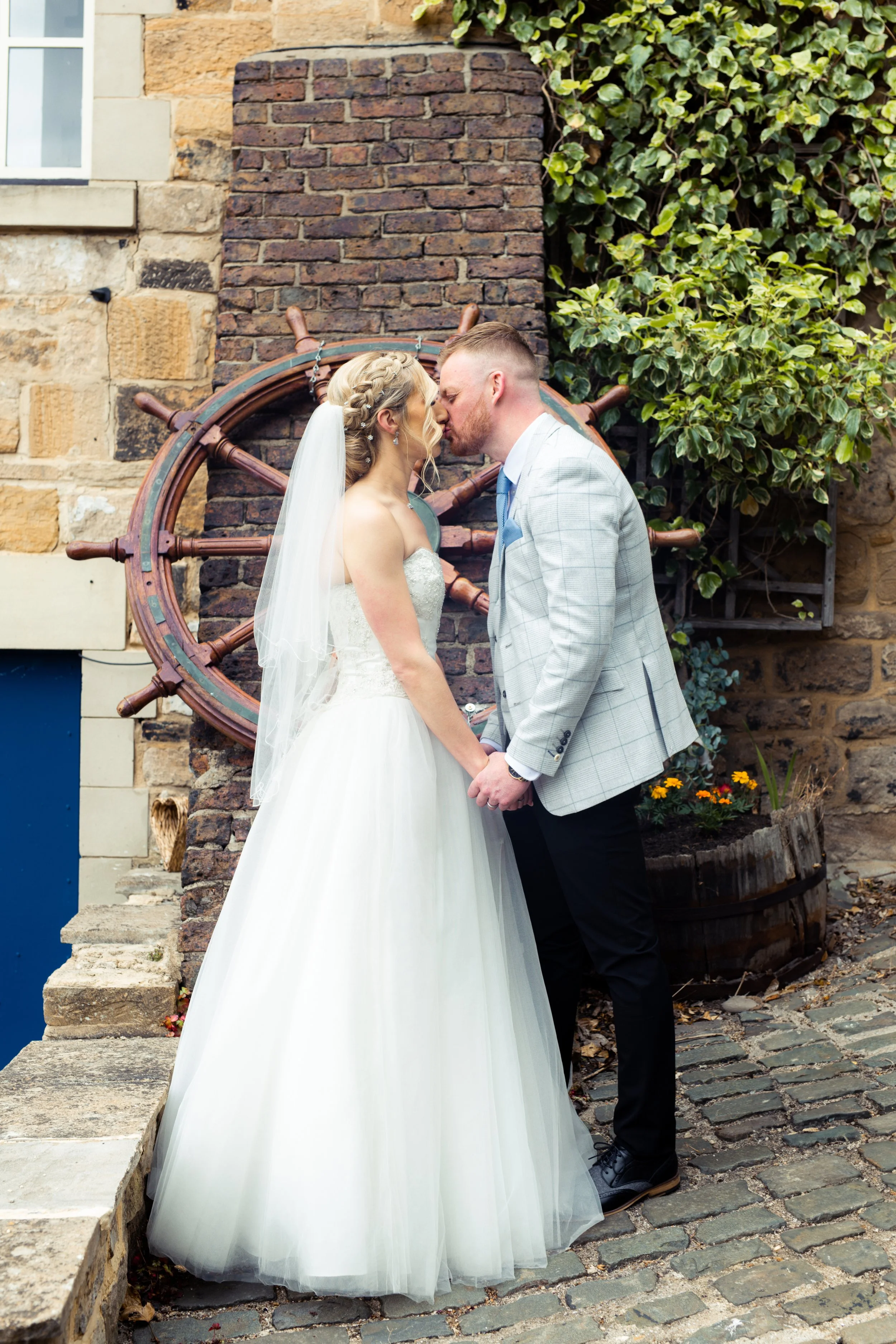 Bride and groom holding hands with foreheads touching, standing on cobblestone street in front of brick wall and ship's wheel, with greenery and flowers in the background.