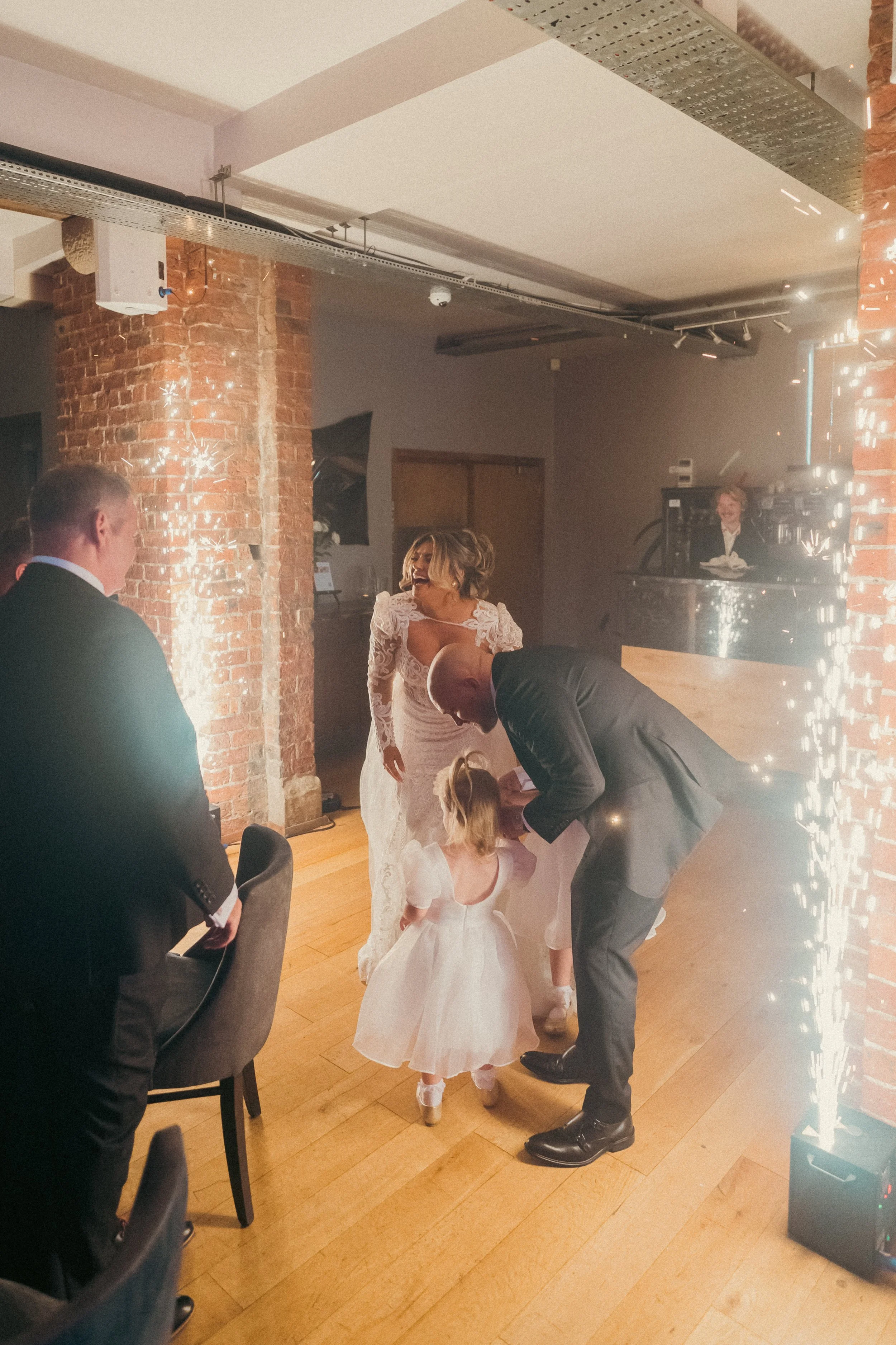 At a wedding reception, a bride in a lace wedding gown and a man in a suit are interacting with two young girls in white dresses, with guests watching in the background.