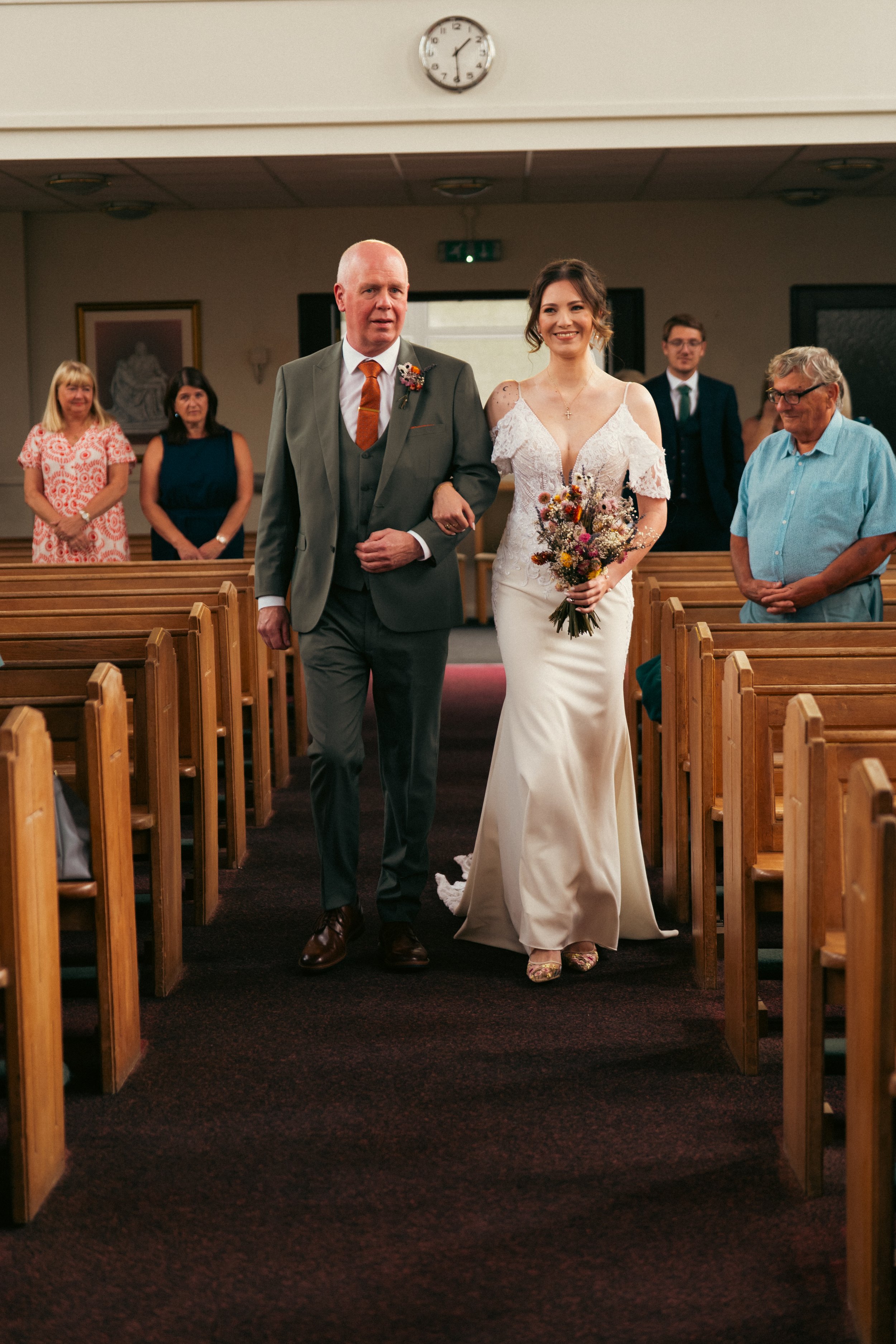 A bride in a white wedding gown walking down the aisle with a man in a gray suit in a church, surrounded by guests.