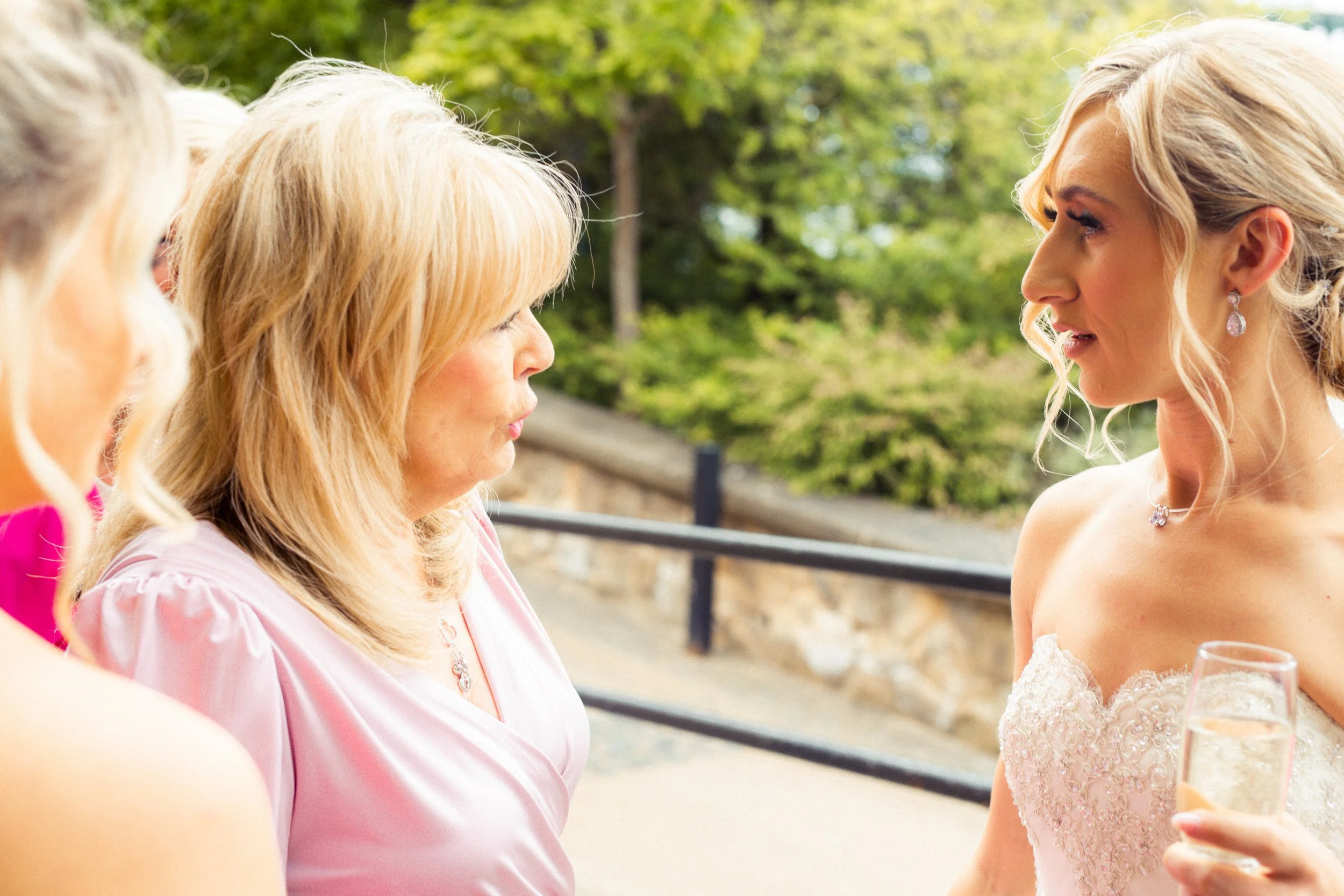 A bride in a wedding dress holding a glass of champagne, conversing with women in pink dresses outdoors with trees and a stone wall in the background.