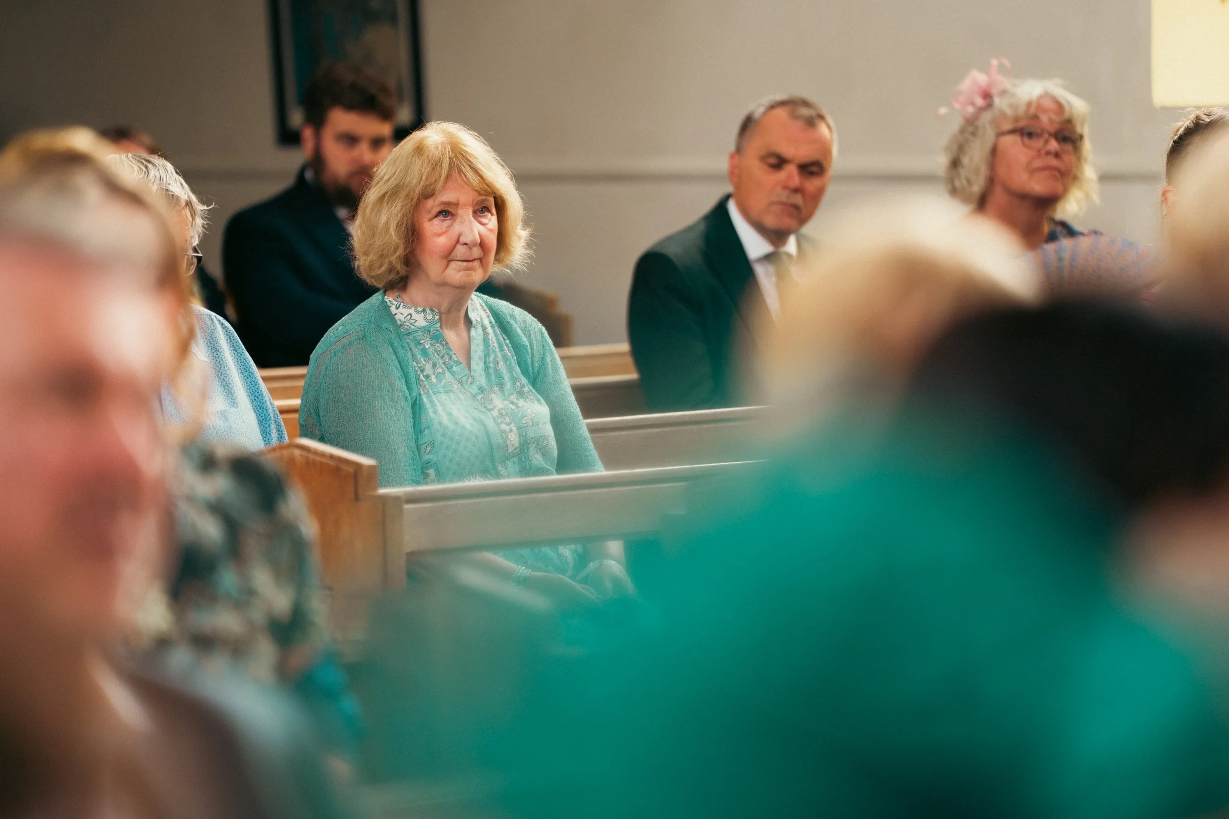 A woman with curly blonde hair wearing a turquoise cardigan and blouse sitting in a church pew during a service. Other attendees are present, some with serious expressions.