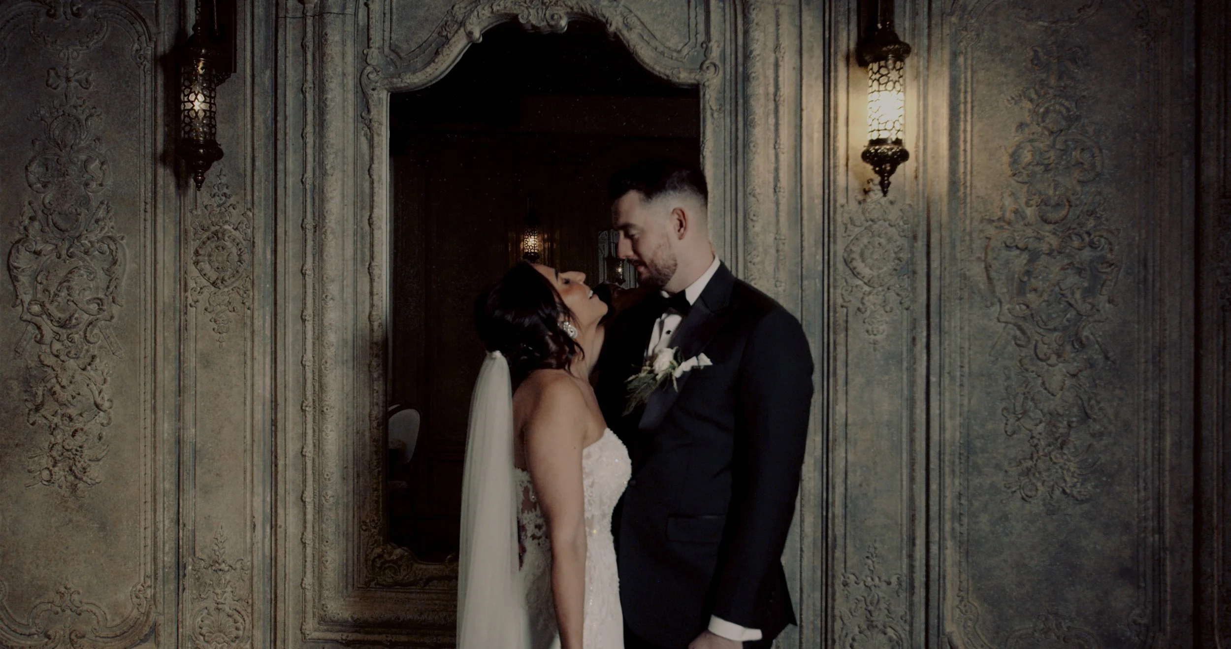 A bride and groom stand close together, gazing into each other's eyes in a decorated, vintage-style room with ornate wallpaper and wall sconces.