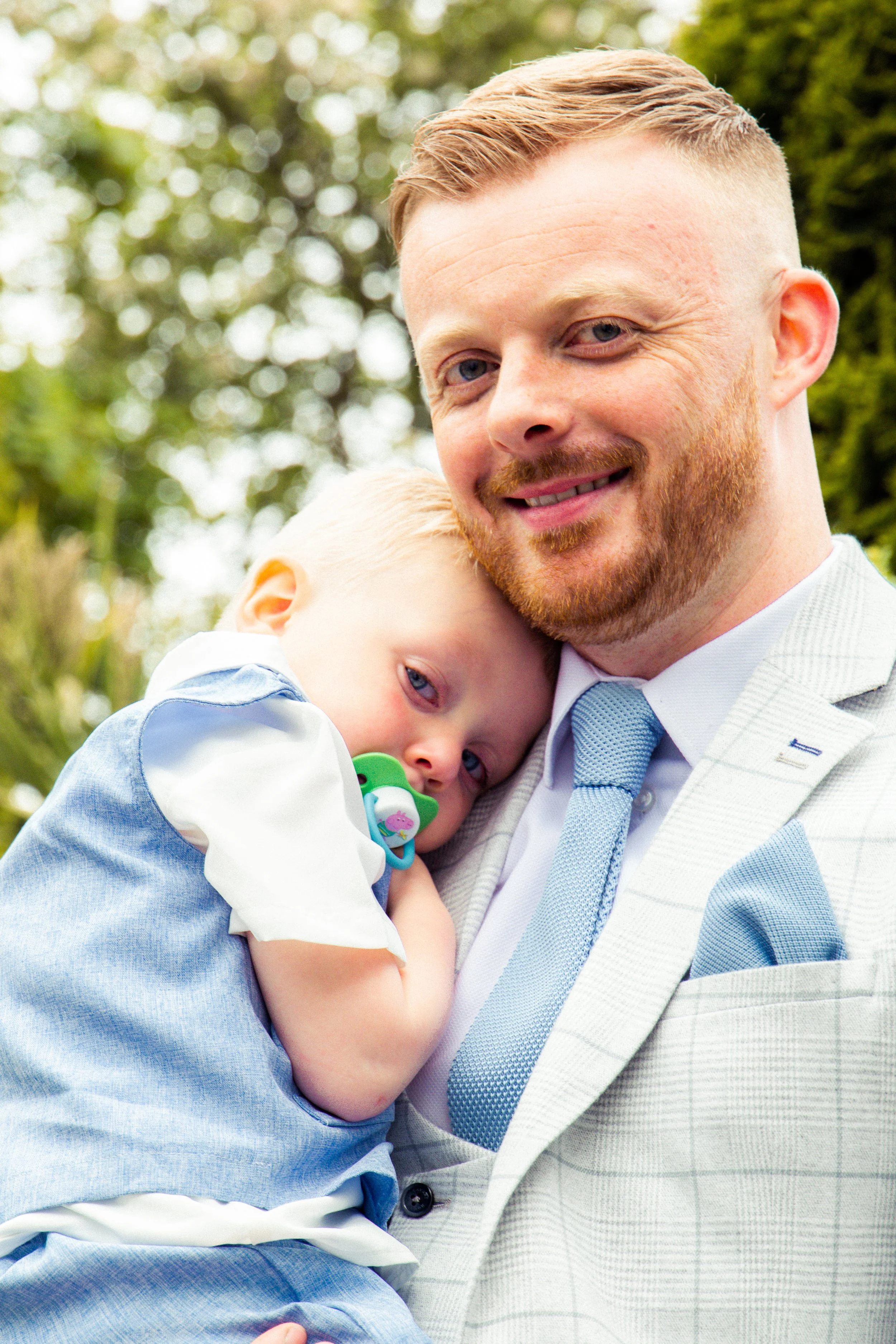 A man with red hair and beard holding a young blond boy with a pacifier in an outdoor setting with green trees in the background.