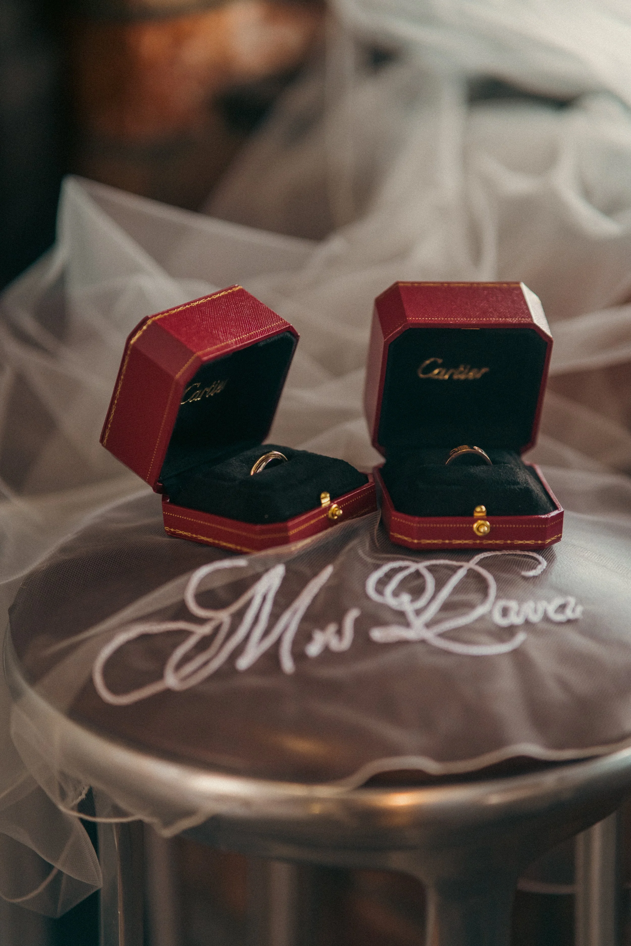 Two small red Cartier jewelry boxes with gold rings inside, placed on a silver tray covered with sheer fabric embroidered with 'Ms. Dana'.