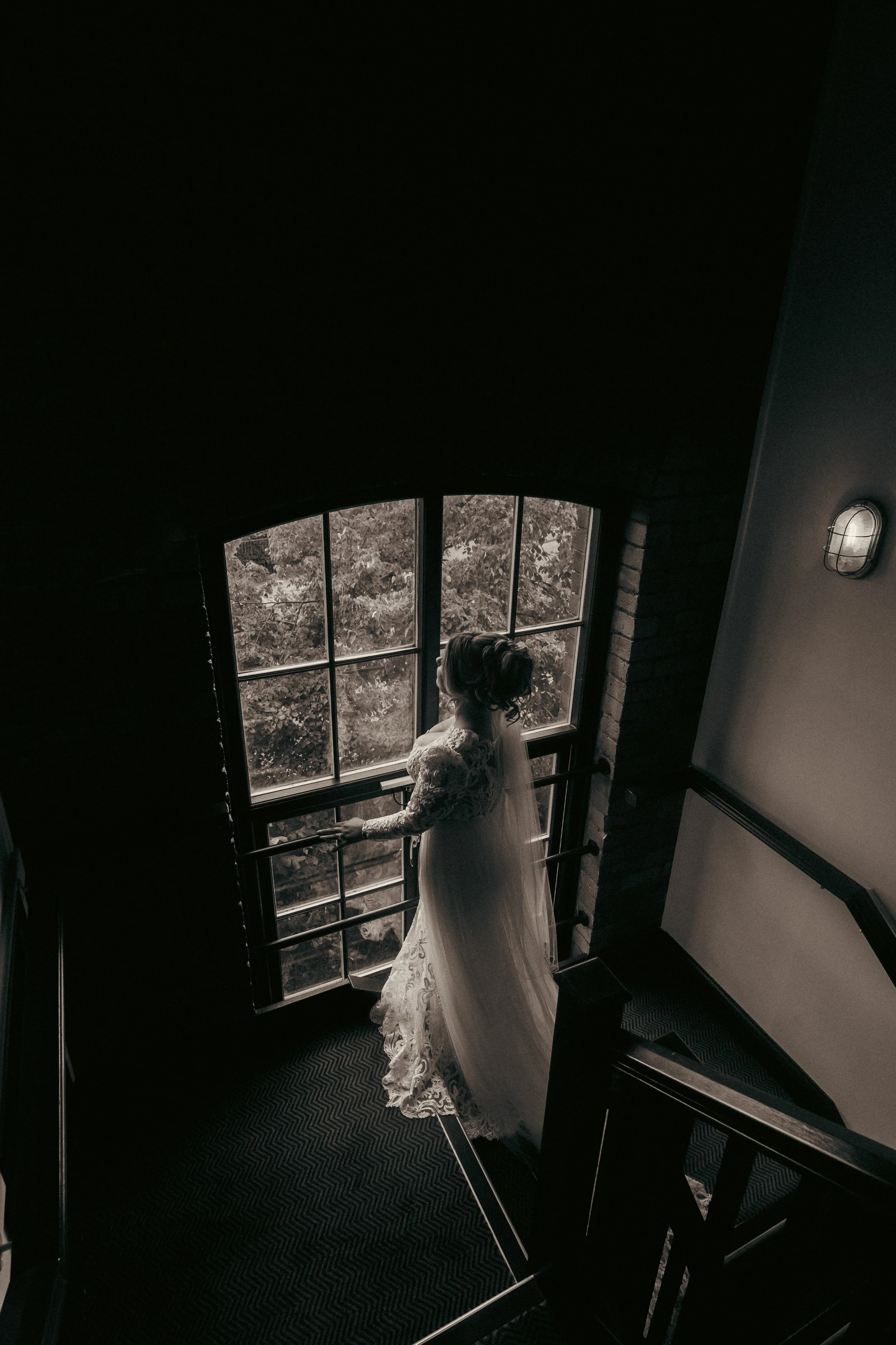 A woman in a wedding dress standing on a staircase, gazing out a large window with trees outside, in a dimly lit indoor setting.