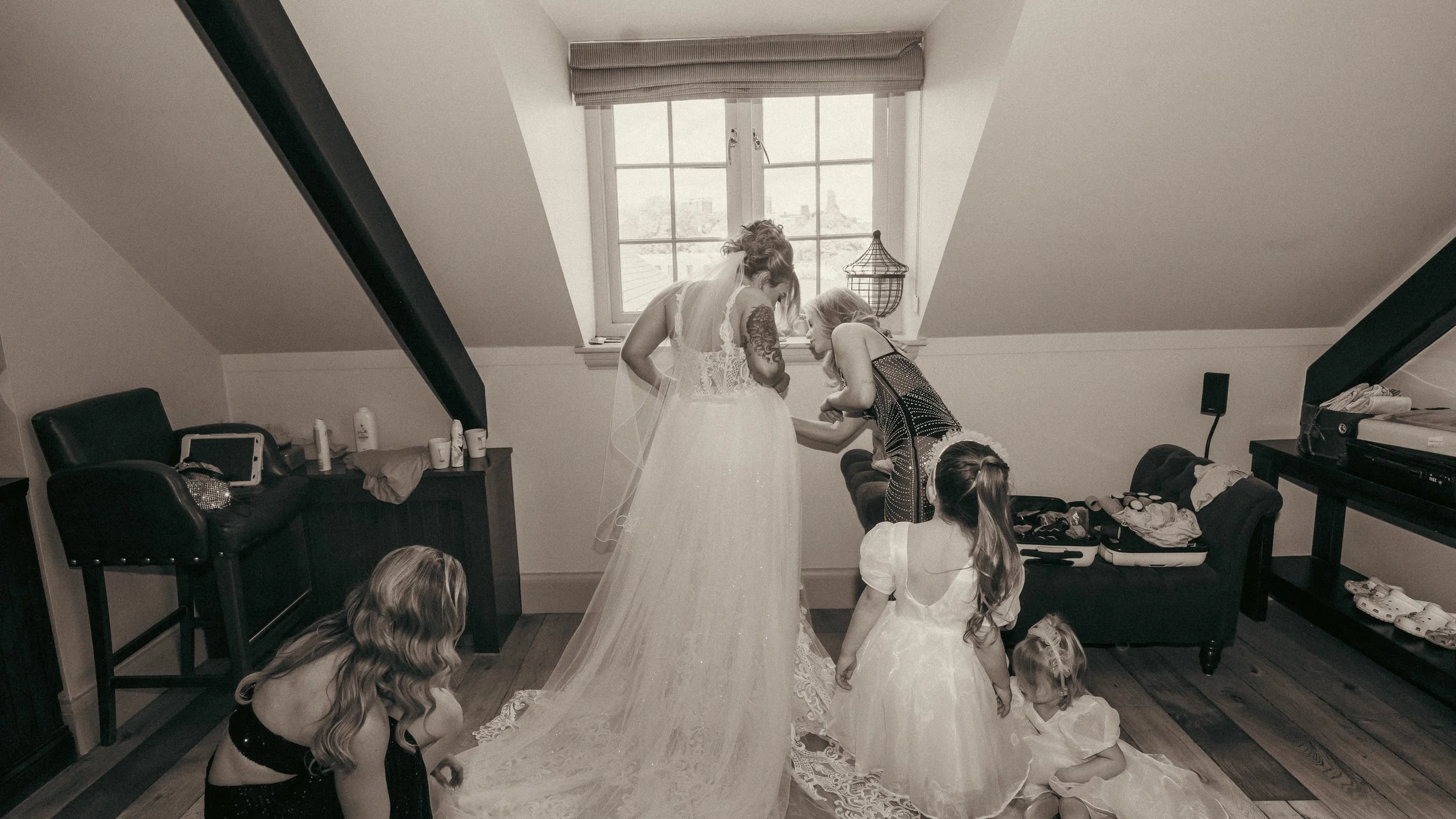 A bride in a wedding gown being assisted with her dress by a woman, with children and onlookers in a bedroom with a large window, black furniture, and various personal items.