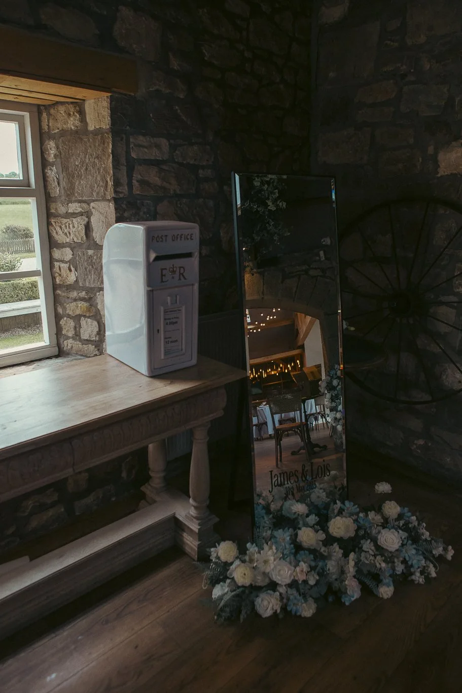 A wedding or event decor setup featuring a mirror with a wedding message, floral arrangements, a stone wall, a wooden table with a letter box, and a window showing an outdoor view.