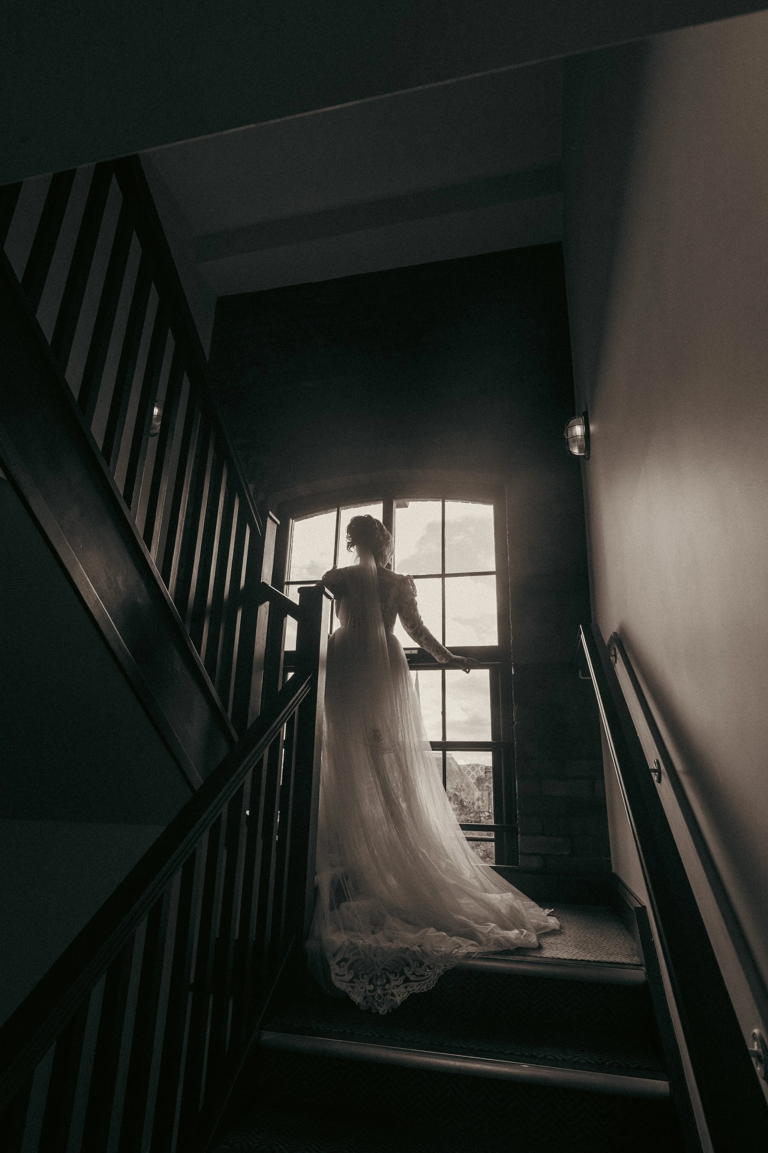 A woman in a wedding dress looks out a large window at the top of a staircase.