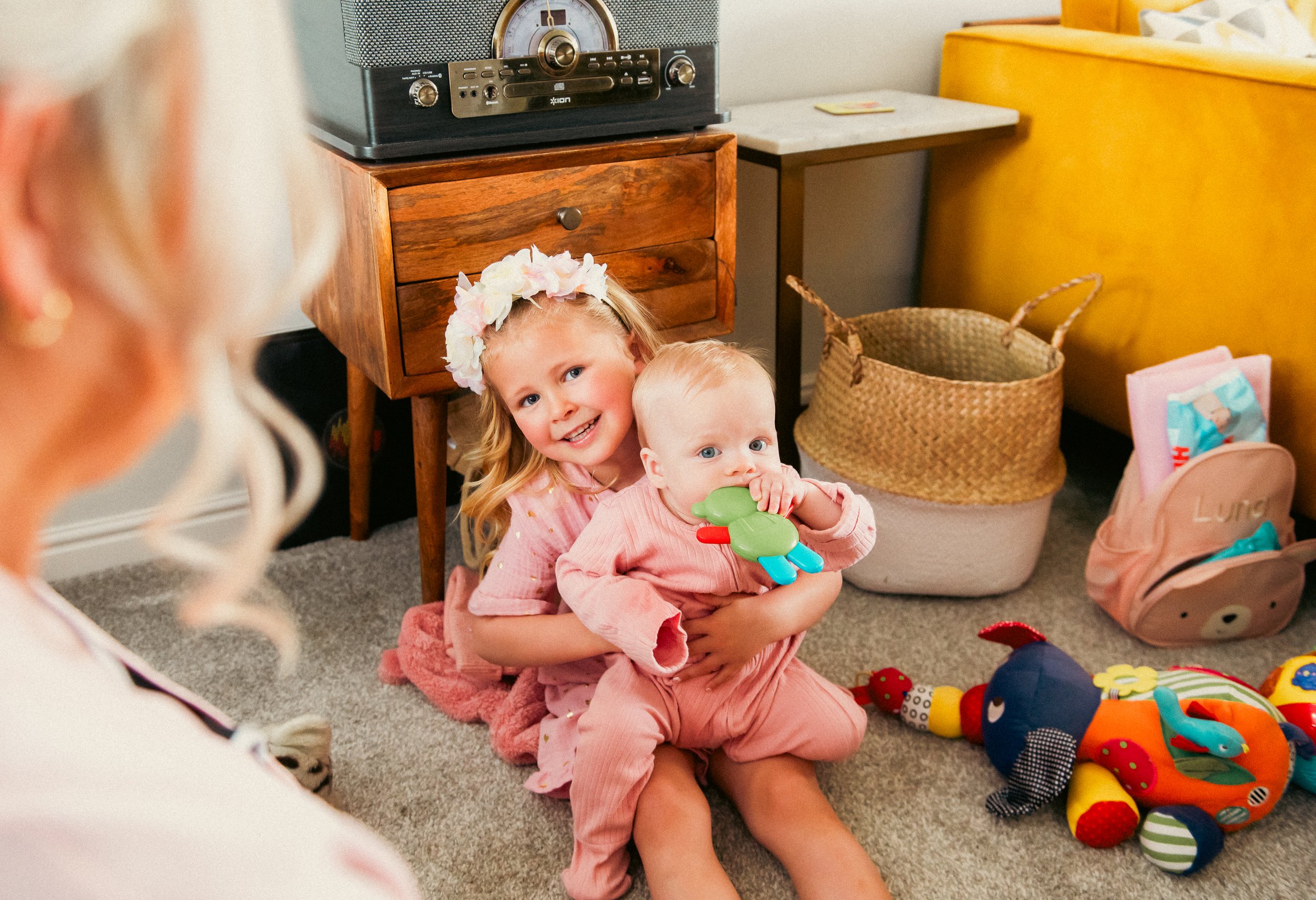 A young girl with a floral headband and a baby holding a green bird toy, sitting on carpeted floor with toys and a woman partially visible in the foreground.