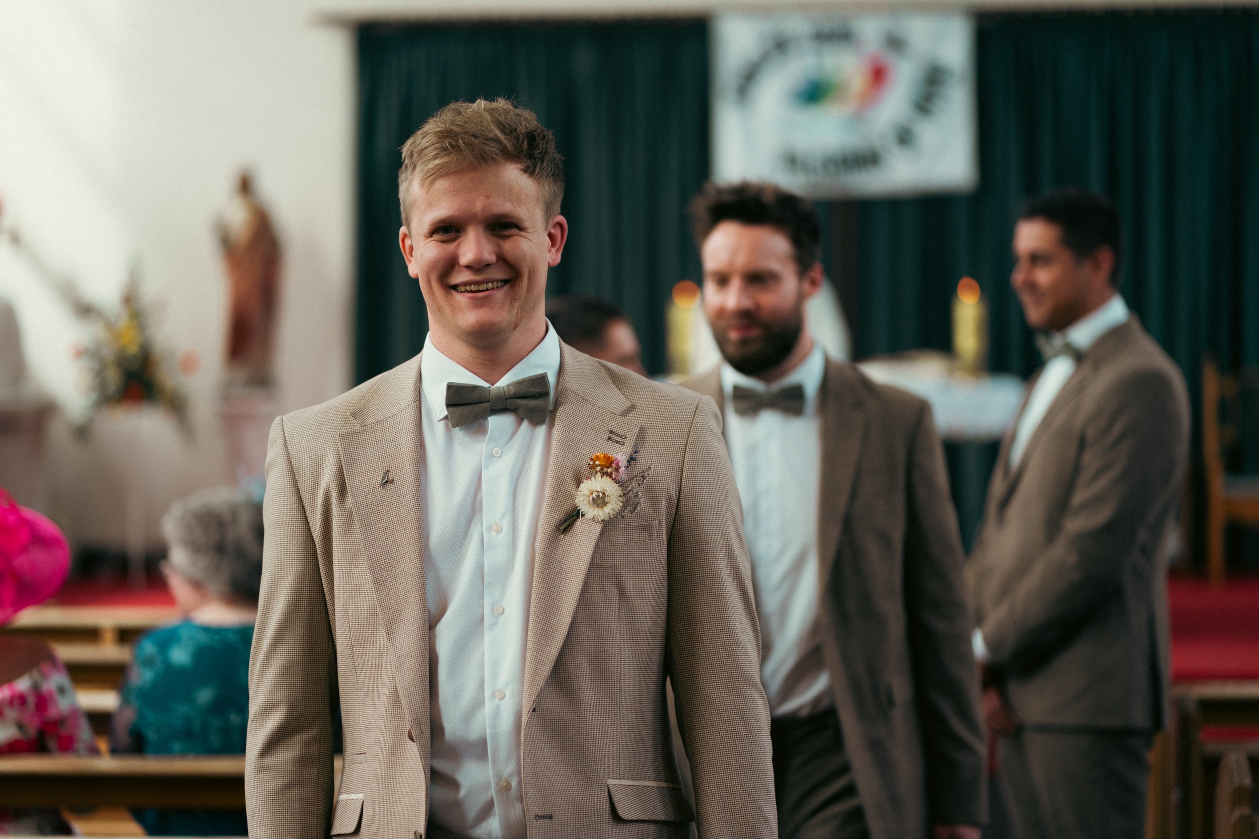 A smiling man in a light beige suit with a bow tie, standing in front of three other men in suits at what appears to be a wedding or formal event, with a green curtain and banner in the background.