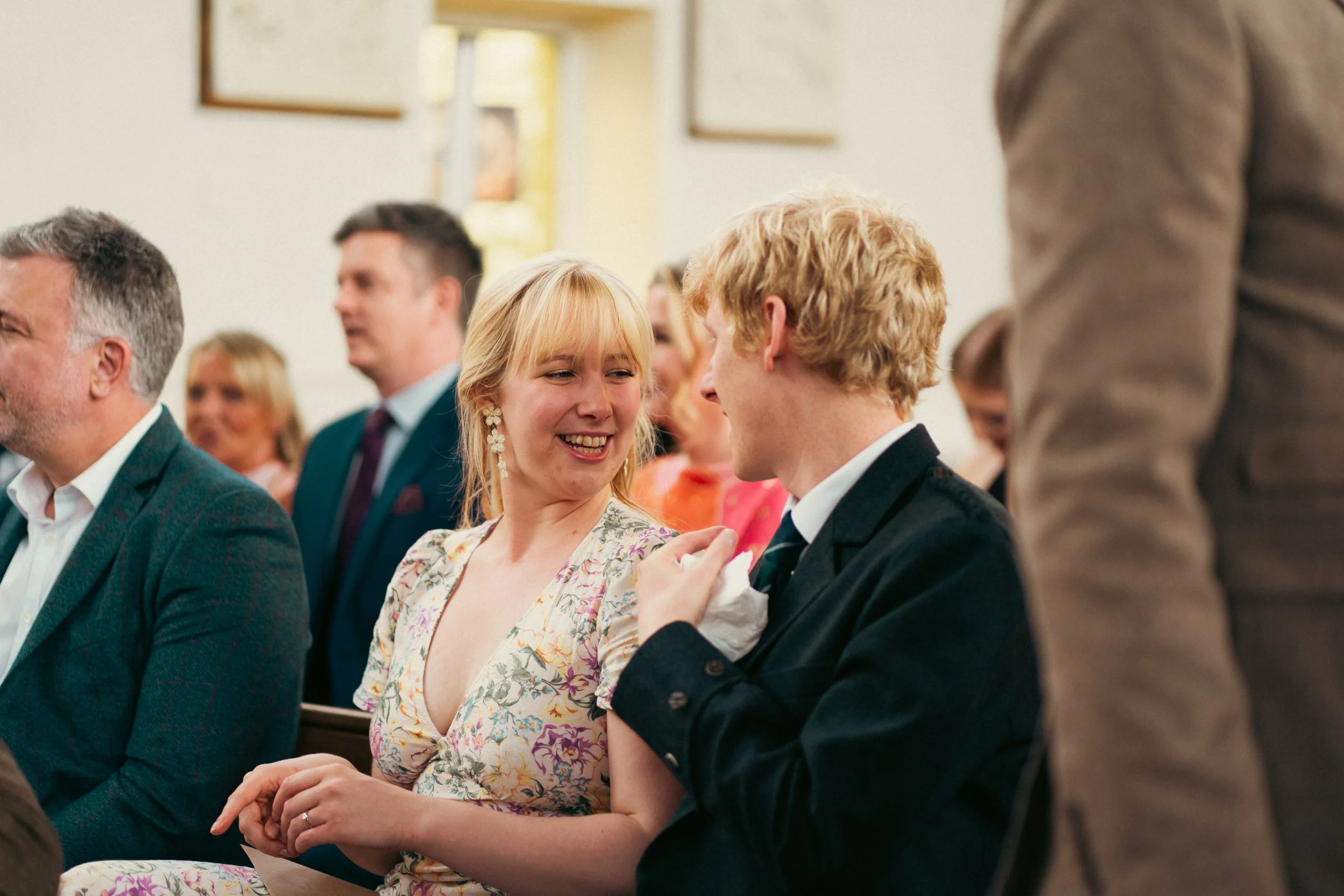A woman and a man sitting next to each other, smiling and engaging in conversation during a formal event, with other attendees seated around them.