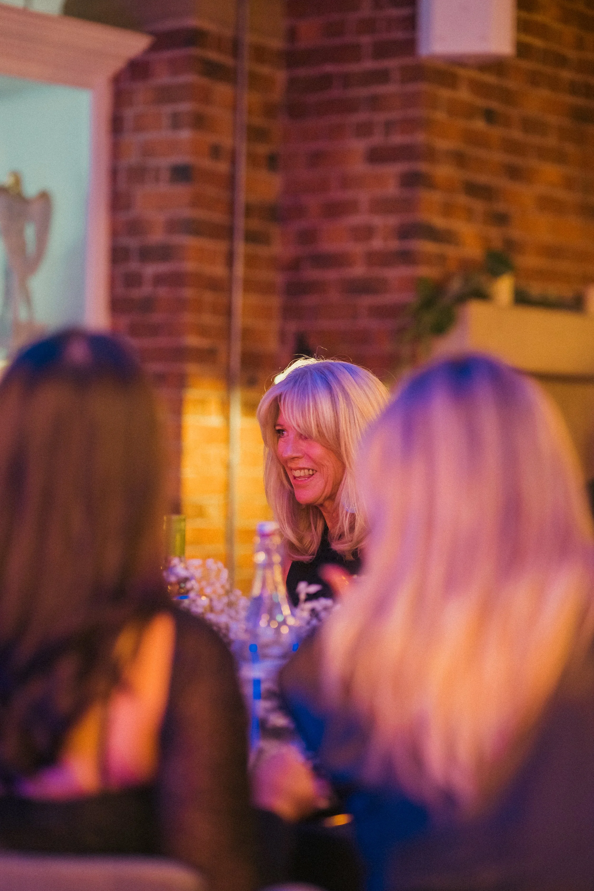 A woman with long blonde hair smiling at a social gathering at a restaurant or bar, with a brick wall background and blurred people in the foreground.
