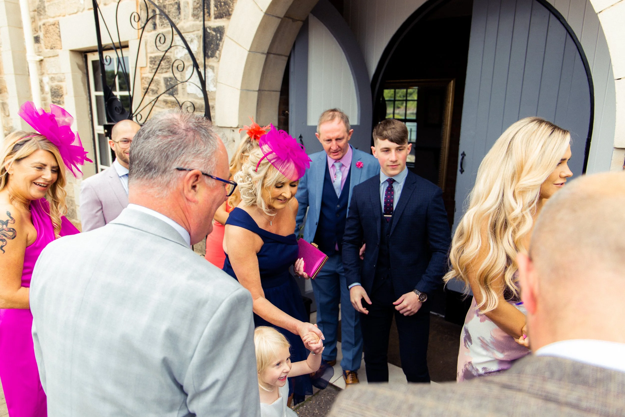 Group of people gathered outside a building, dressed in formal attire, with some wearing bright pink and purple hats, during a celebration or wedding.