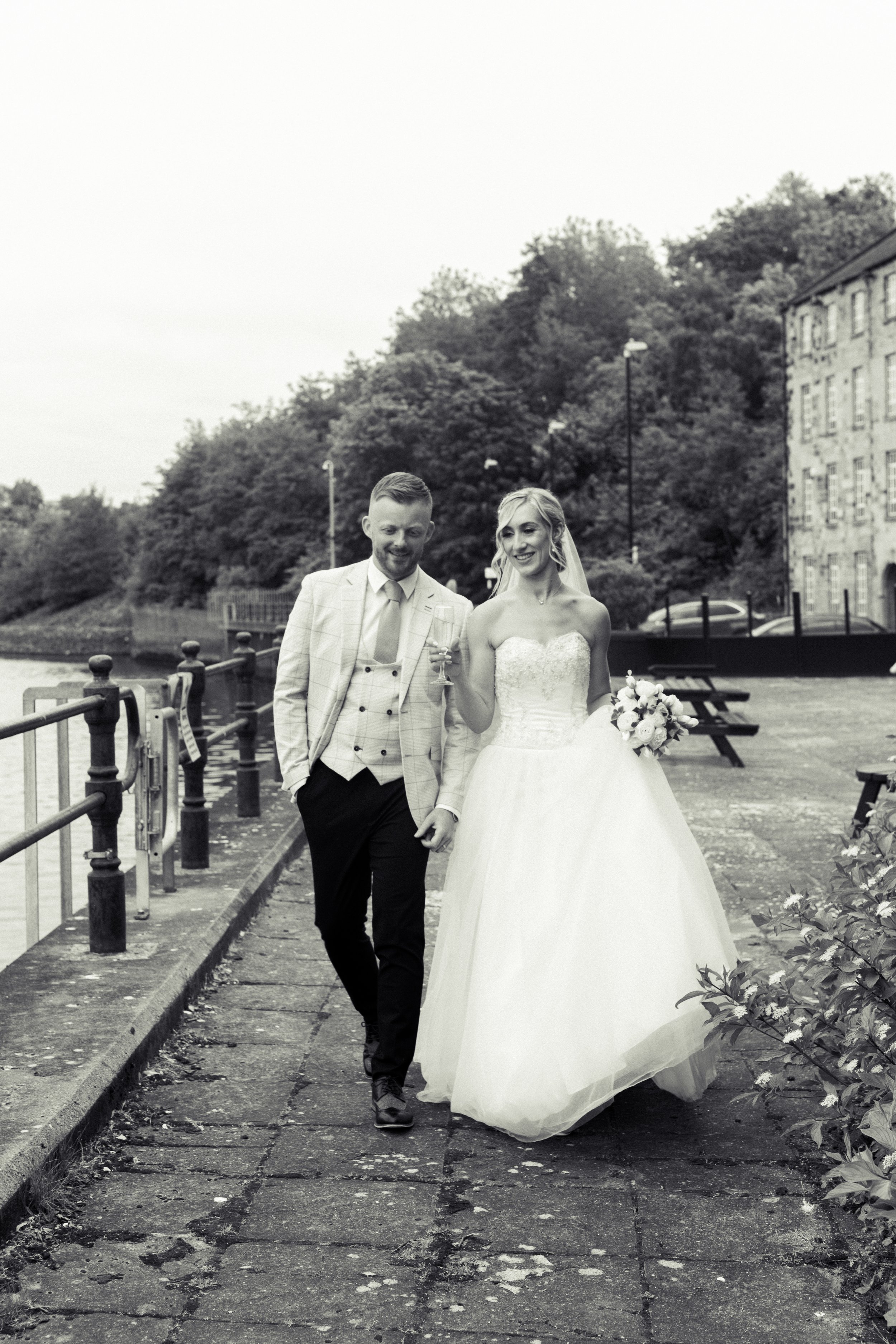 A bride and groom walking along a riverside path, smiling and holding a champagne glass, with trees and buildings in the background.