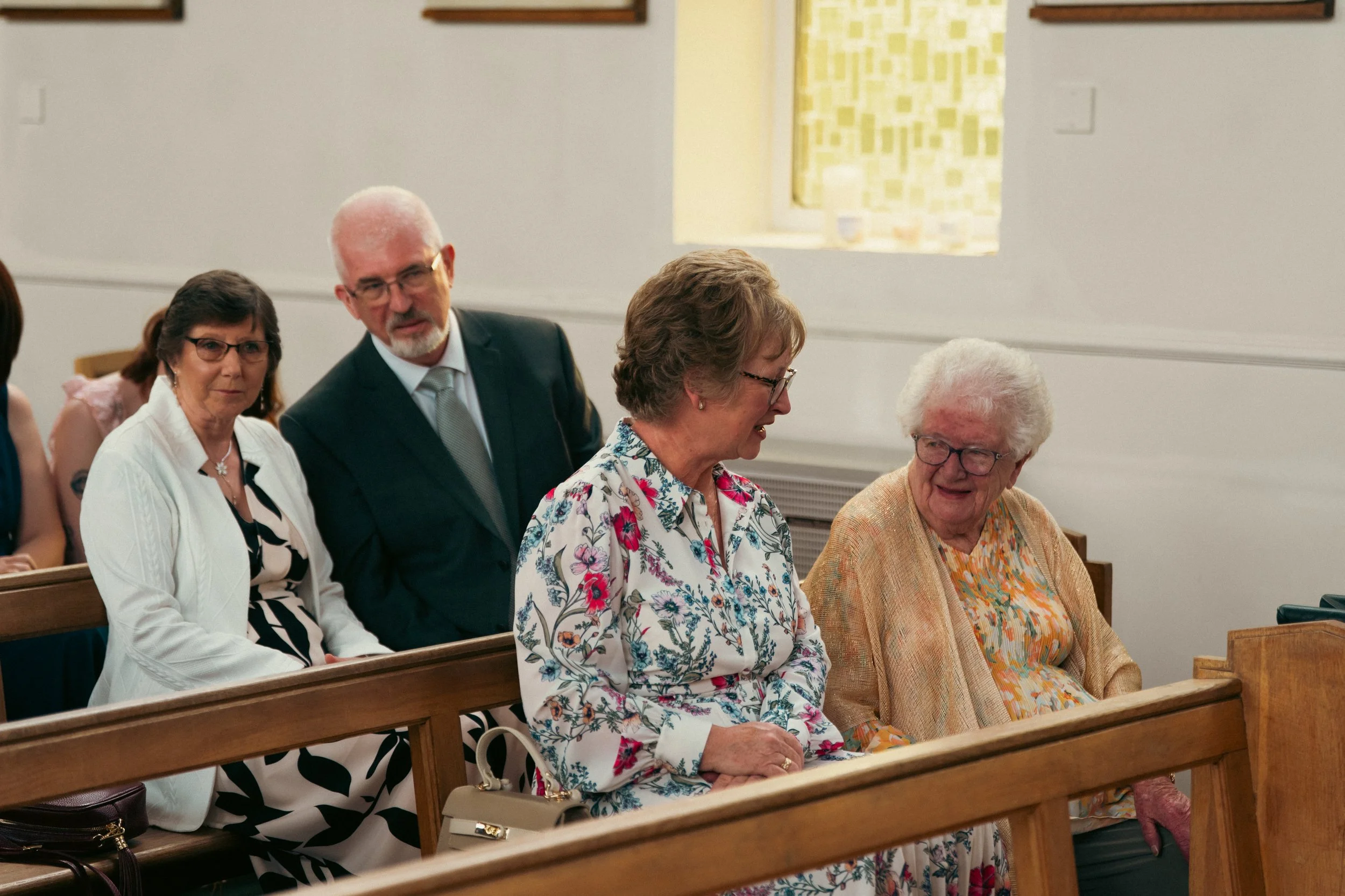 Group of elderly women and one elderly man sitting and standing inside a church pew, engaging in conversation during a gathering or service.