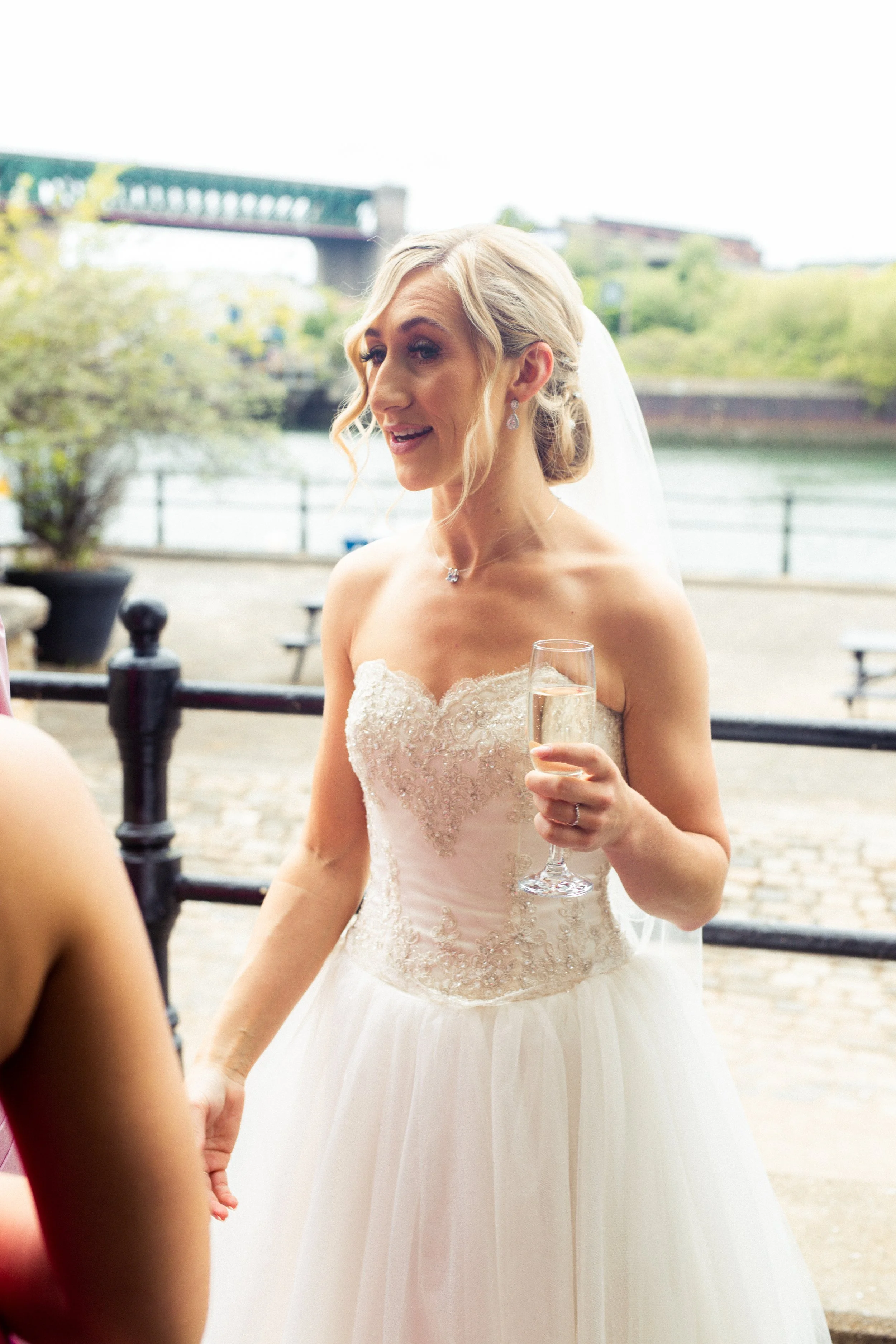 A bride in a strapless lace wedding gown holding a glass of champagne, smiling outdoors near a river with a bridge in the background.