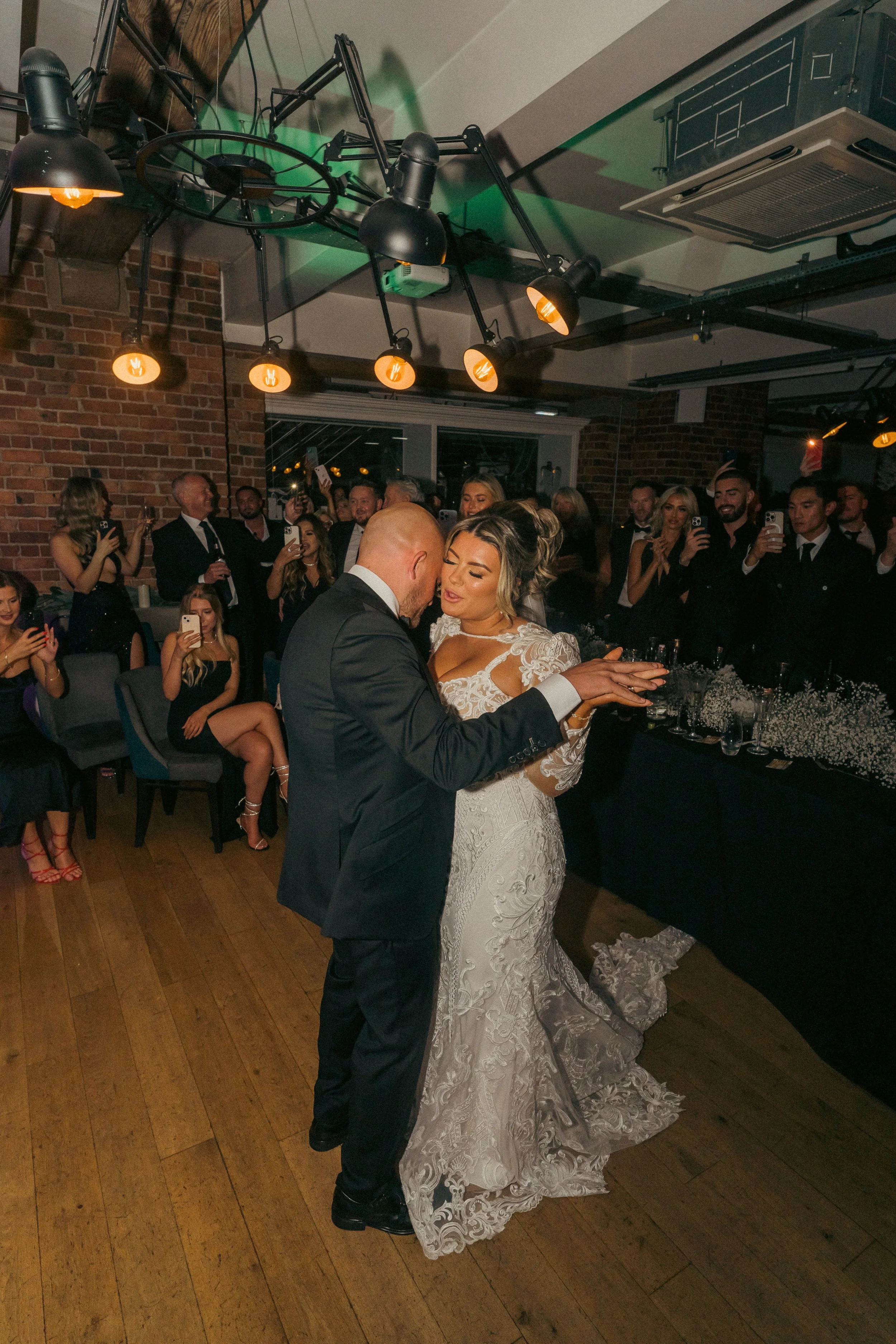 A bride and groom share a dance during their wedding reception, surrounded by guests taking photos and watching in a dimly lit room with brick walls and industrial ceiling fixtures.