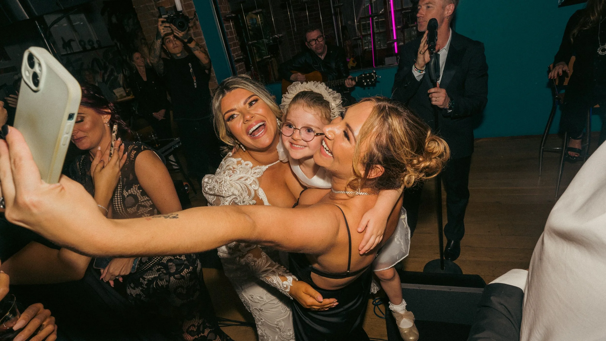 Group of women and a young girl taking a selfie at a celebratory event, with a singer performing in the background.