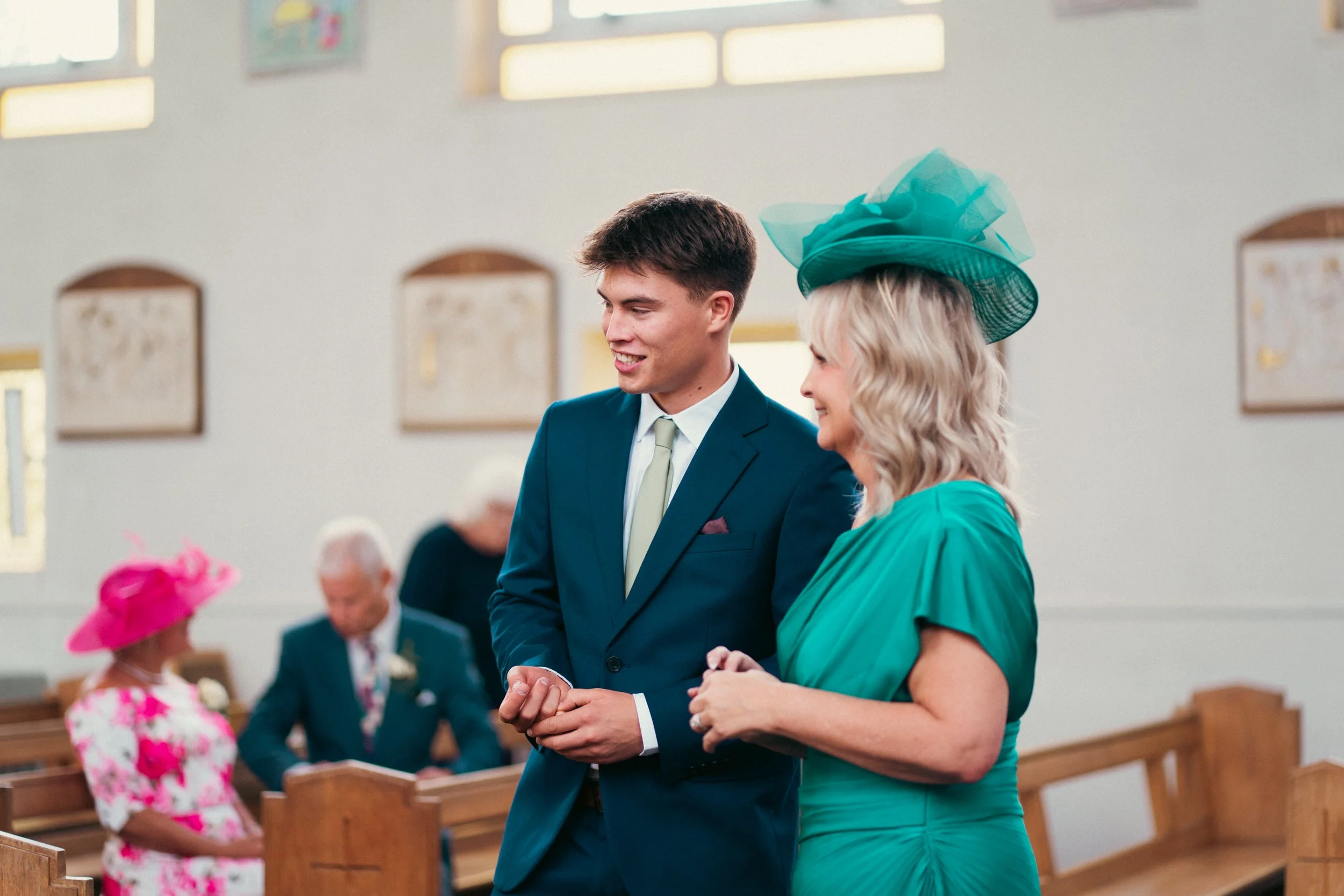 A young man in a blue suit and a woman in a teal dress with matching hat are holding hands and smiling inside a church. In the background, an older man, an older woman, and a girl wearing a pink hat and dress are present.