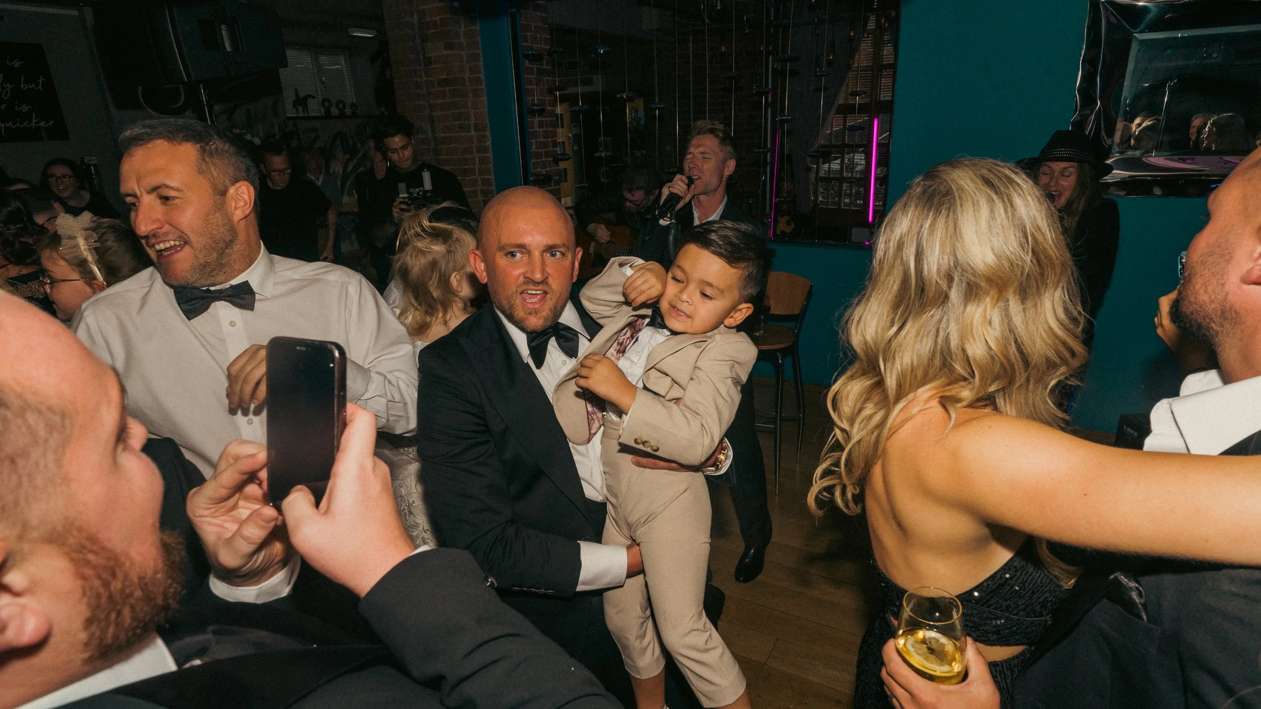 People dancing and celebrating at a party, some dressed in formal attire like suits and tuxedos, in a lively indoor setting.