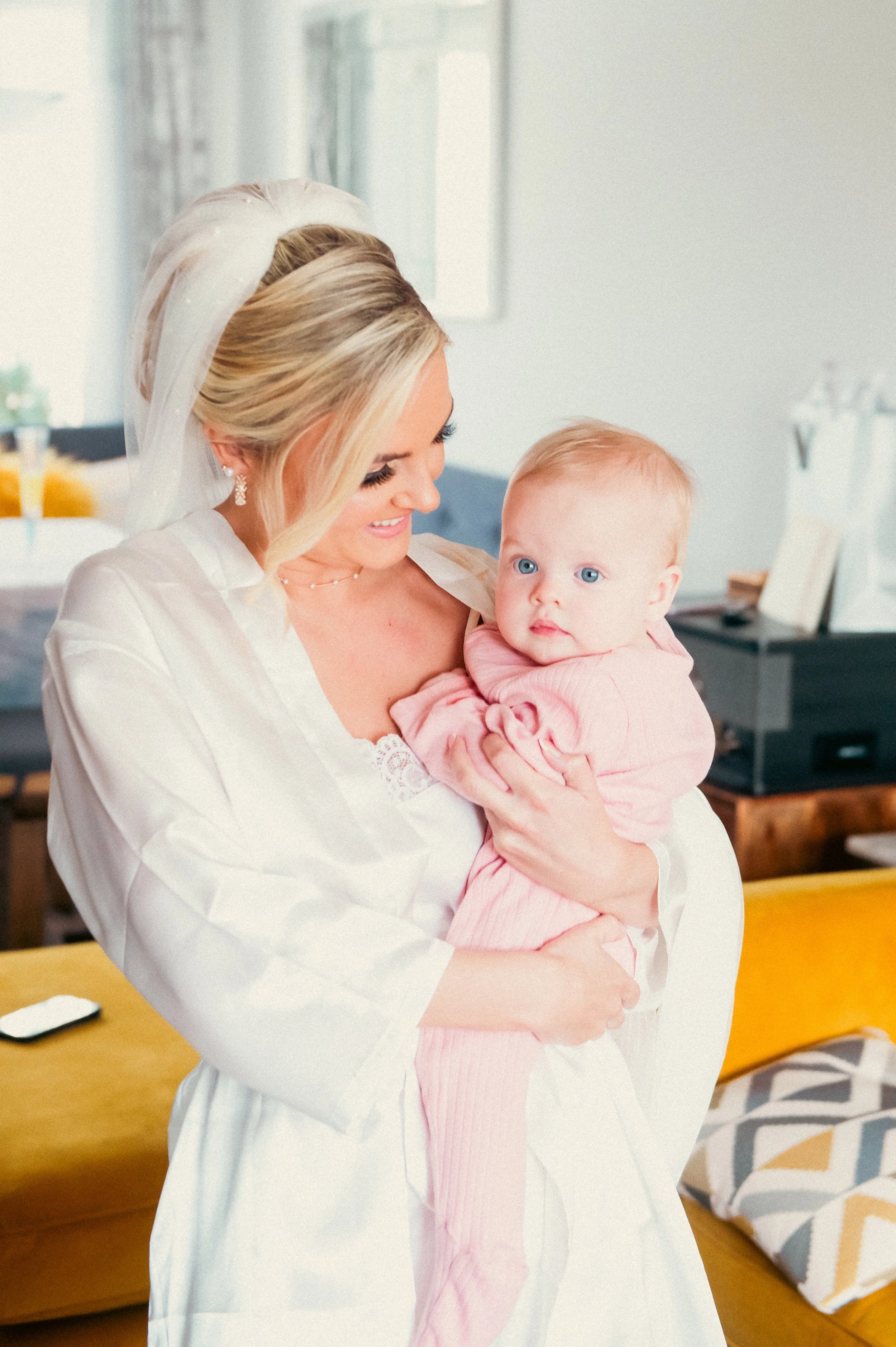 A woman dressed as a bride holding a young child in a pink outfit indoors.