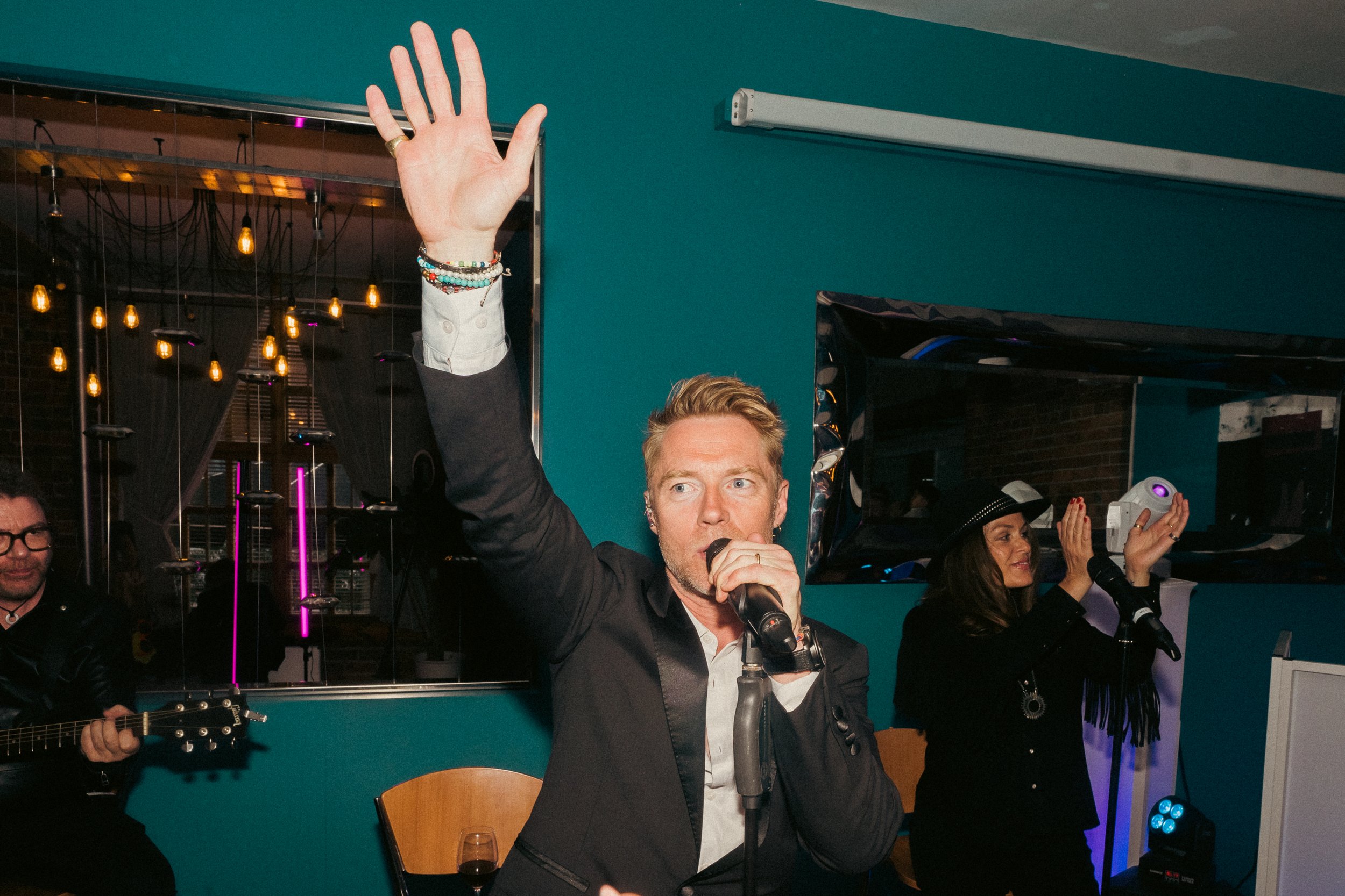 A man with blonde hair singing into a microphone with a raised hand, performing at an indoor event with musicians and audience members nearby.