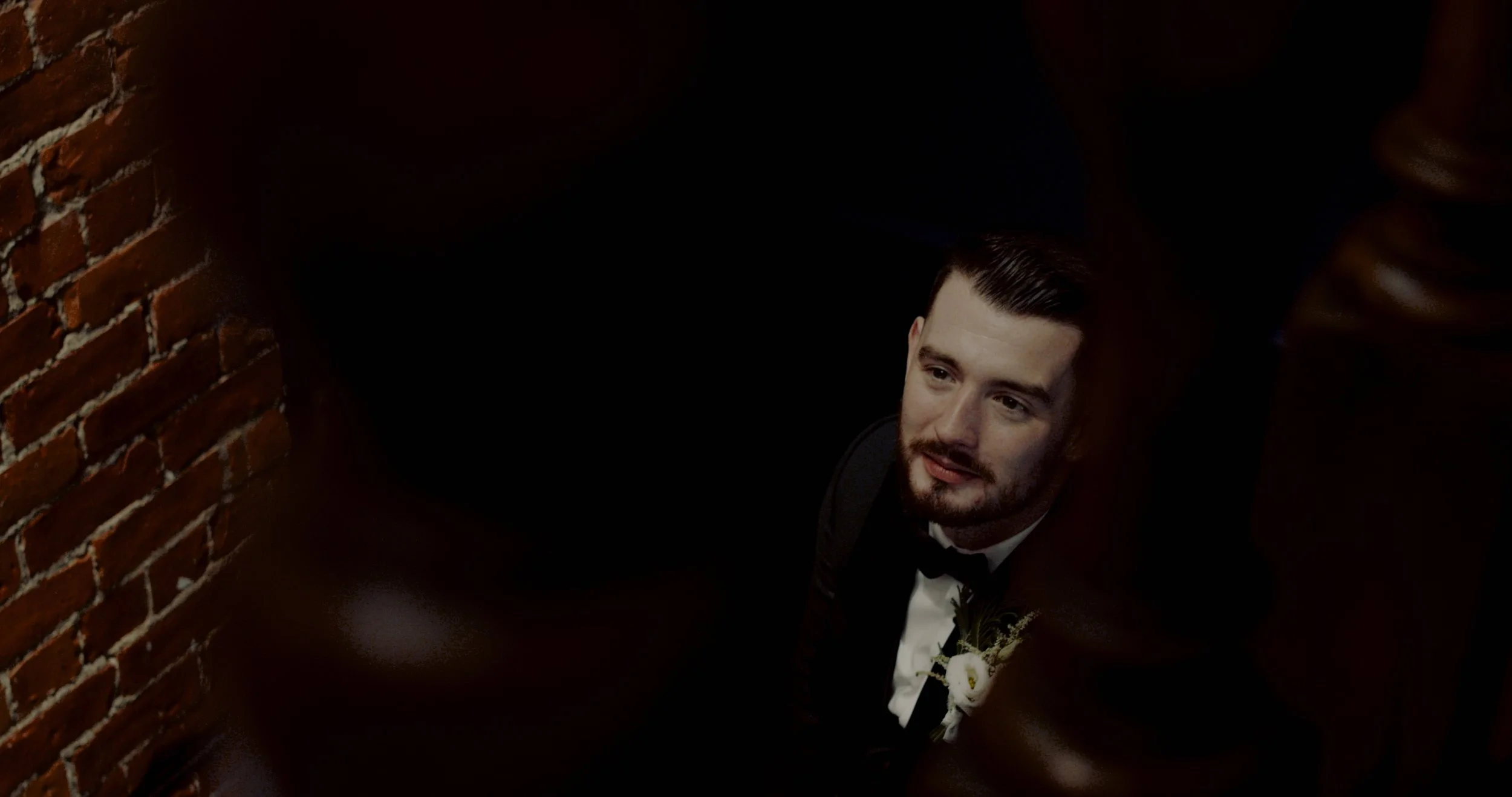 A man dressed in a tuxedo with a boutonniere, sitting in a dark corner with brick wall behind him, looking up at the camera.