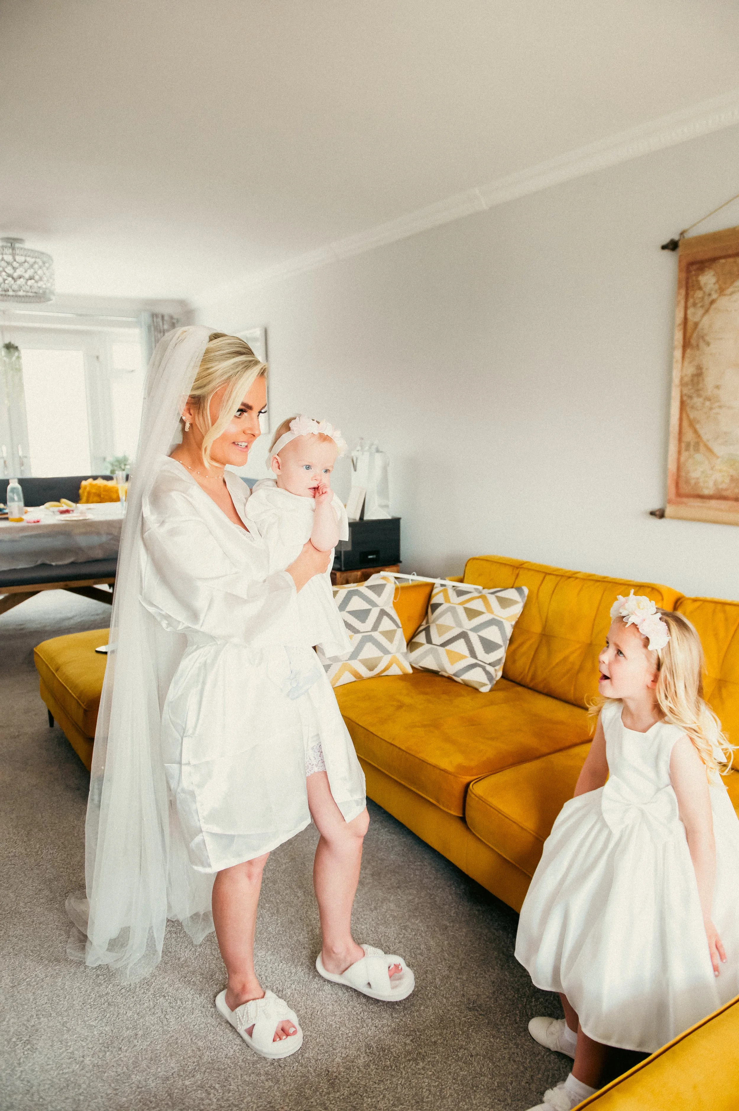 A bride in a white dress and veil holding a young girl in a white dress, standing in a living room with a yellow couch and a girl in a white dress looking up at them.