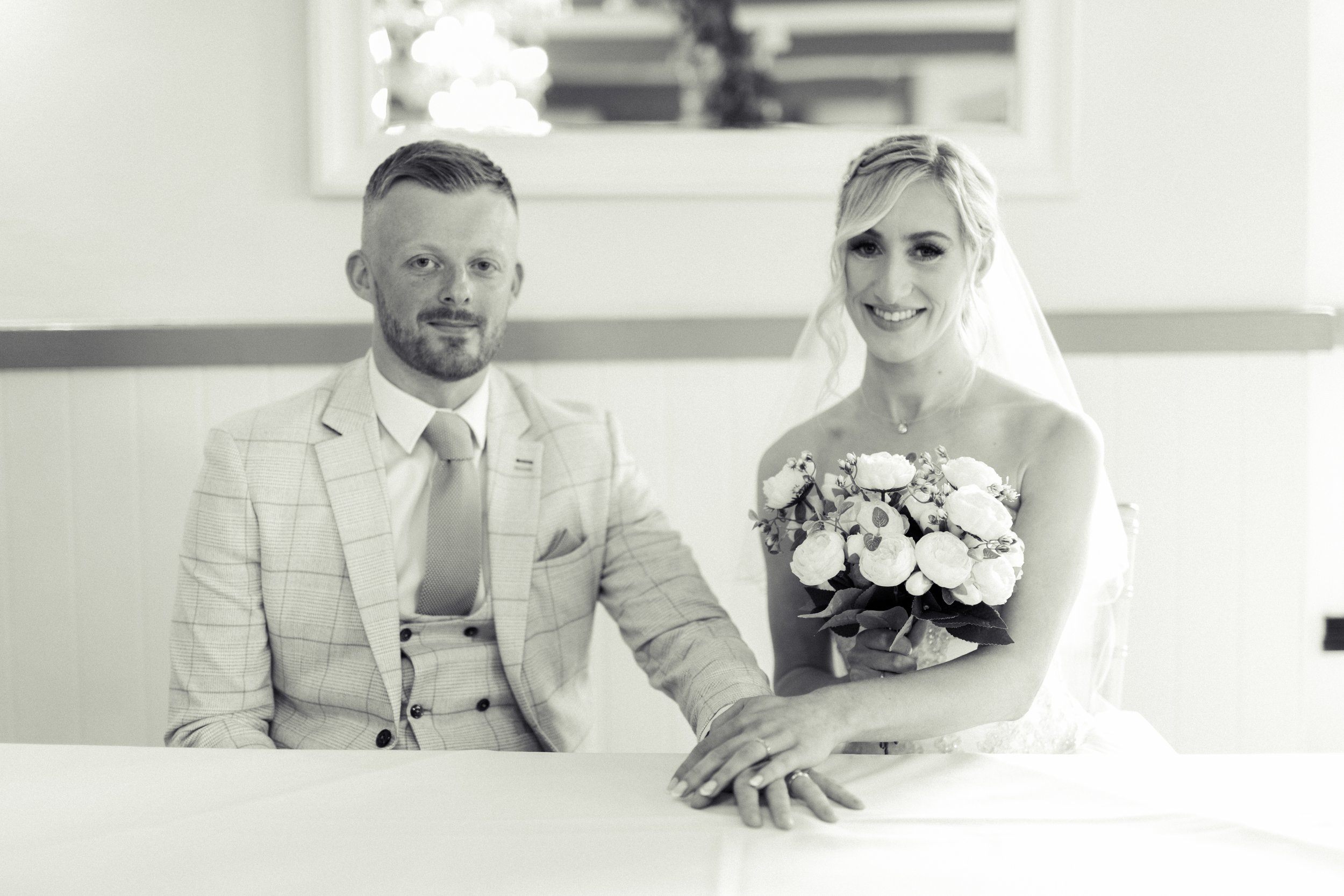 A black and white photo of a bride and groom sitting at a table, holding hands, with the bride holding a bouquet of flowers and smiling at the camera.