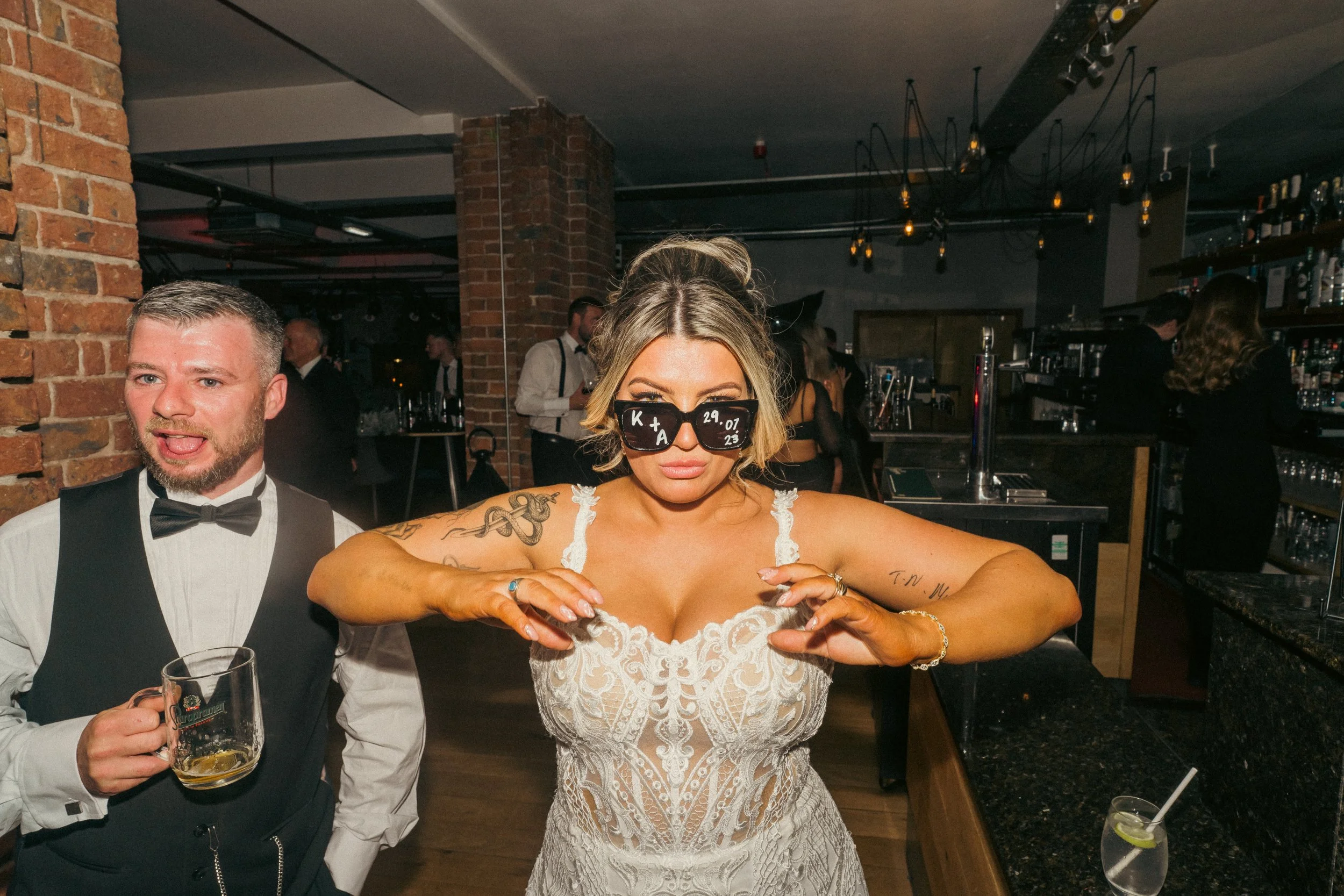 Woman with tattoos wearing a white lace dress and sunglasses, dancing at a bar with bartender and other patrons in background.