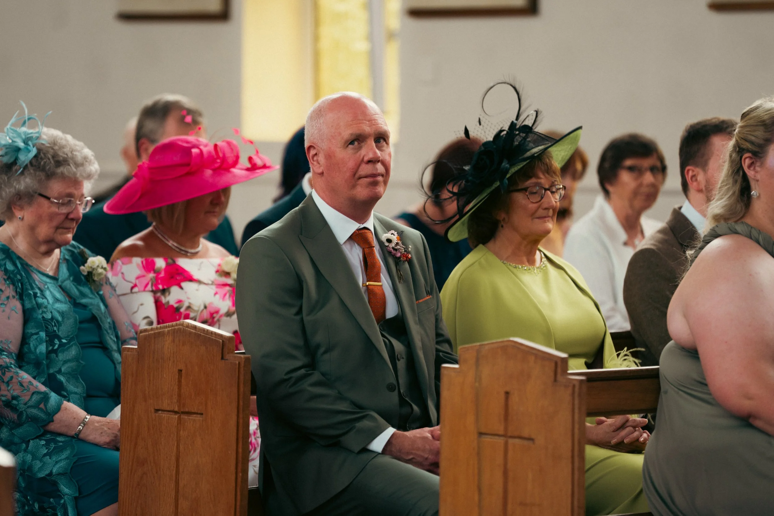 People attending a wedding service seated in a church. Two women wear elaborate hats, one pink and one black, while a man in a gray suit with a boutonniere sits with a woman in a green dress and hat. Other guests are visible in the background.