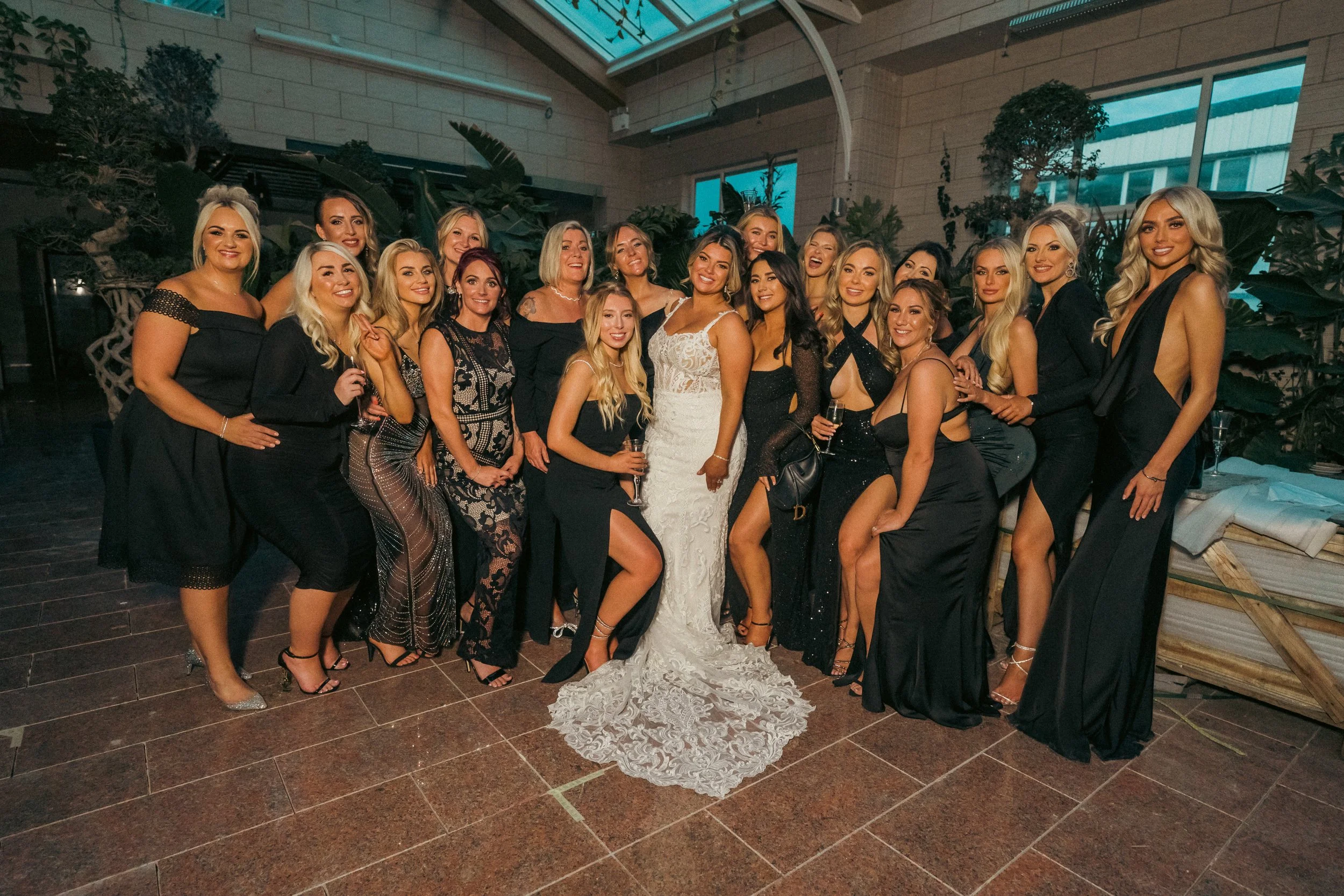 Group of women dressed in formal evening wear, including black dresses and a white wedding gown, posing together at an indoor event with greenery in the background.