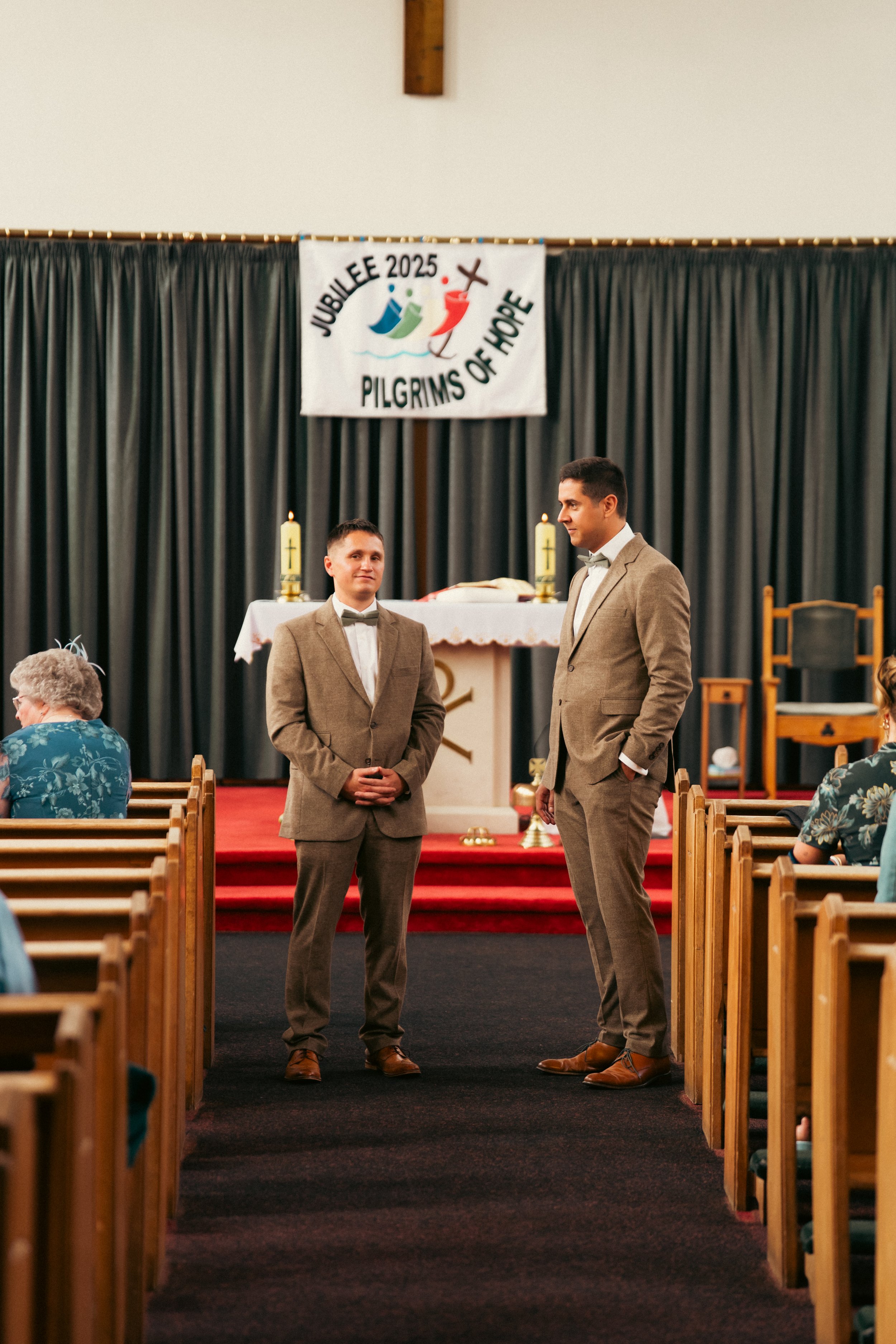 Two men in tan suits standing in front of an altar in a church during a wedding ceremony with a "Jubilee 2025 Pilgrims of Hope" banner hanging above in the background.
