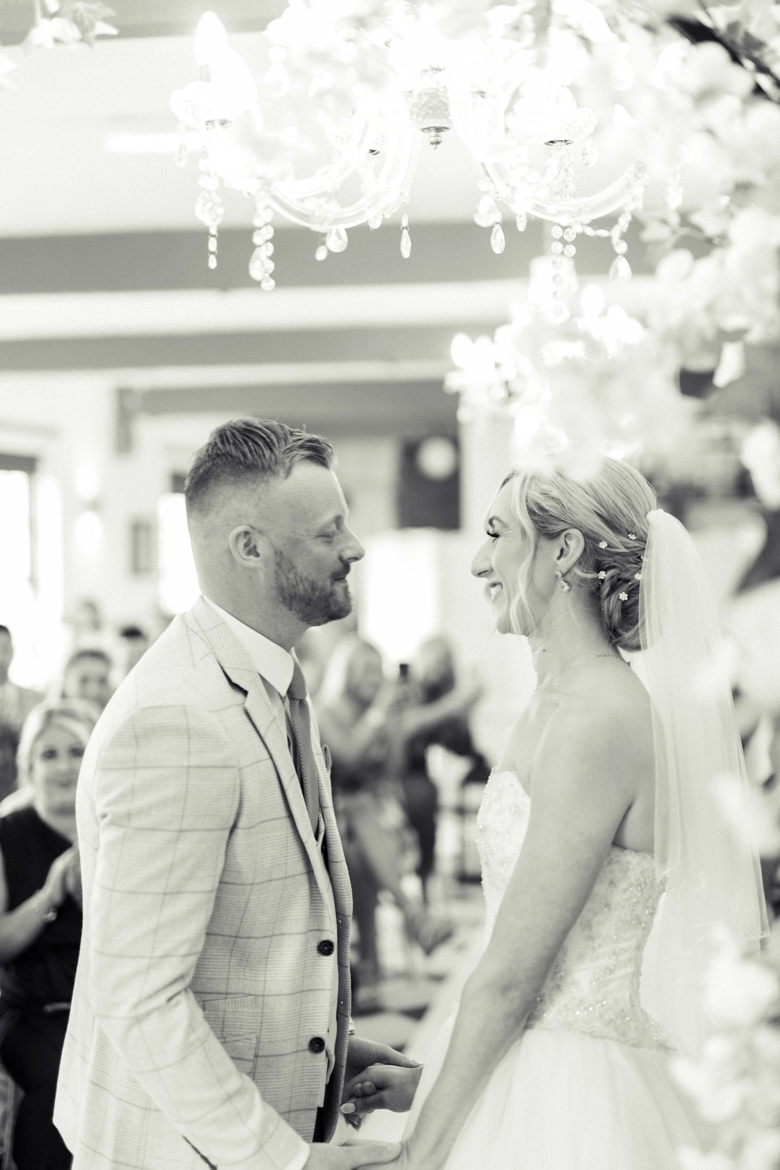 A bride and groom holding hands and smiling at each other during their wedding ceremony in a decorated indoor venue.