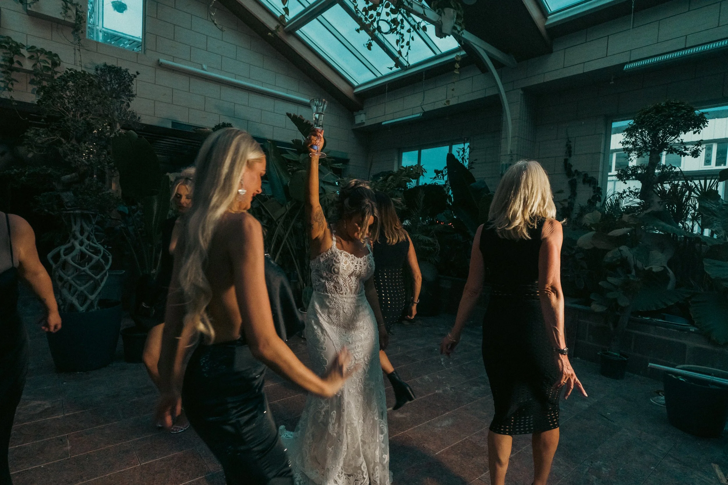 Women dancing in a glass-roofed indoor venue with plants and large windows.