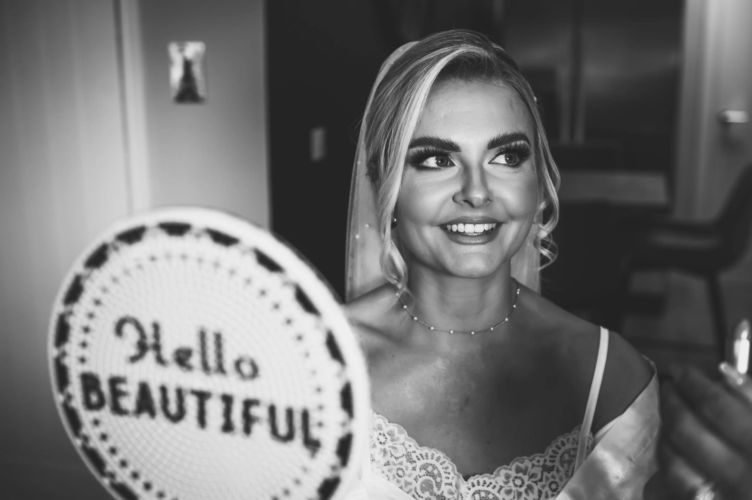 Black and white photo of a smiling woman with a veil, dressed in lacy bridal attire, applying makeup in front of a mirror with a sign that reads 'Hello Beautiful'.