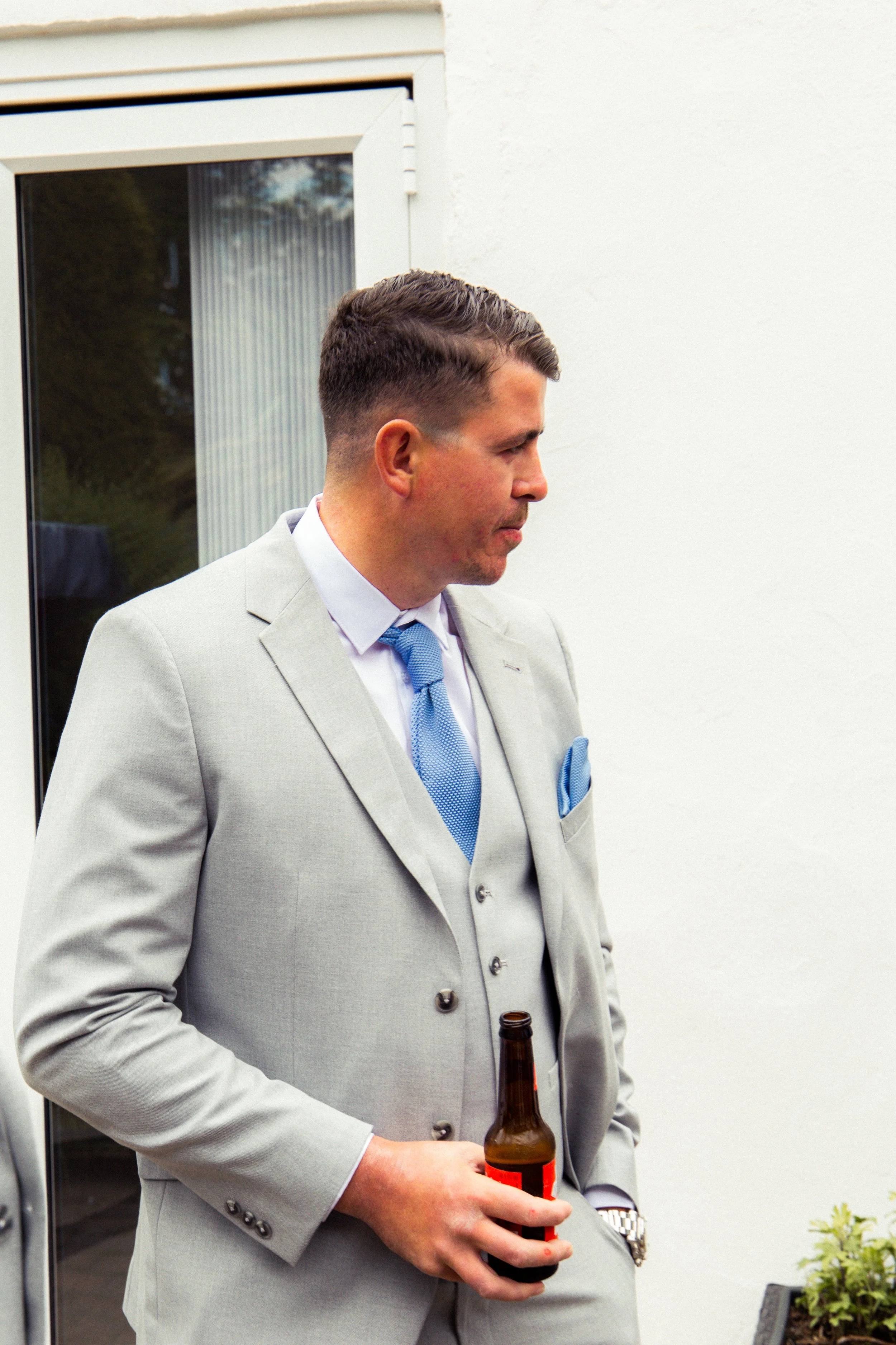 A man in a light gray suit holding a beer bottle, standing outside near a glass door and white wall.
