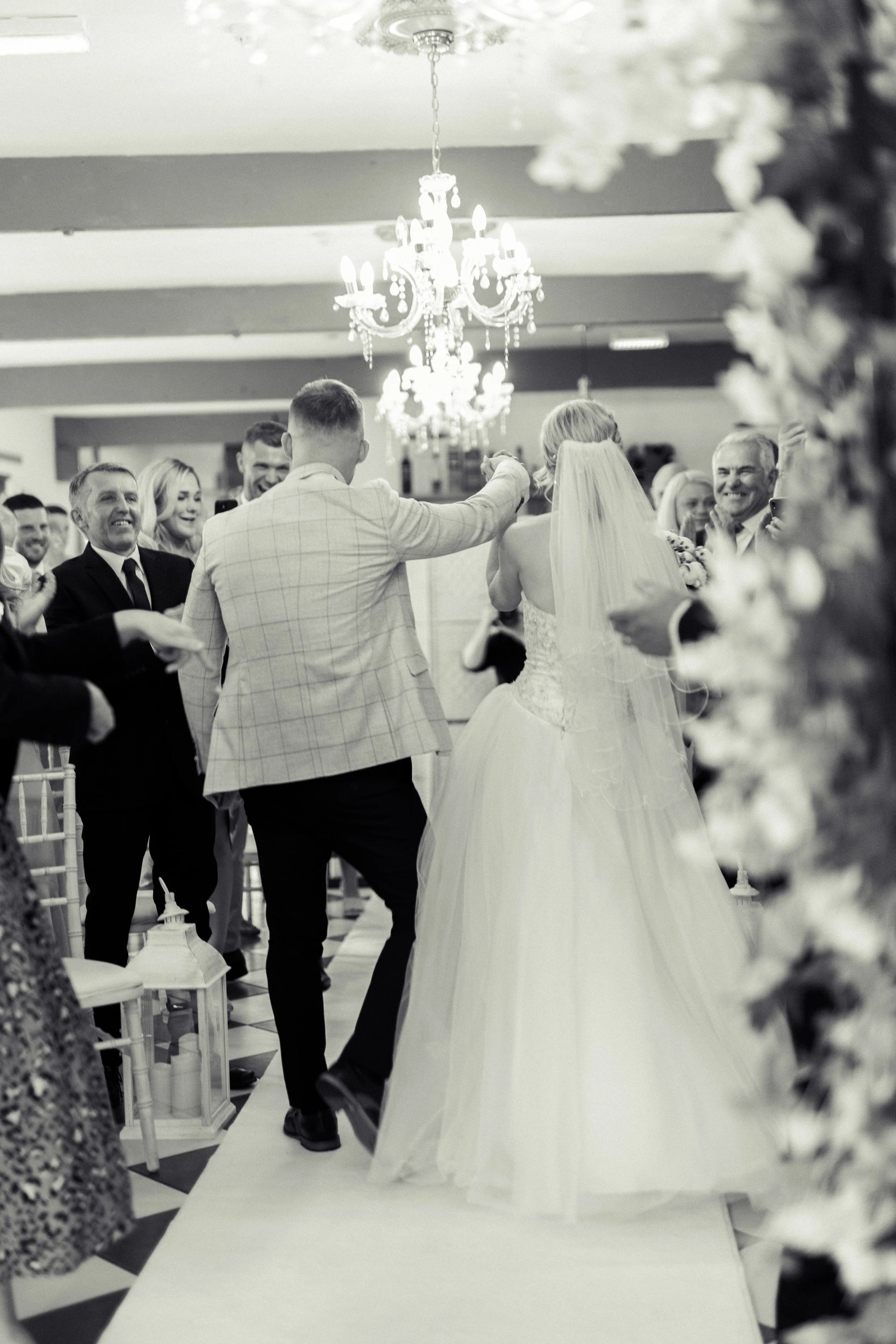 Black and white photo of a wedding reception with a bride and groom dancing under a chandelier, surrounded by smiling guests.