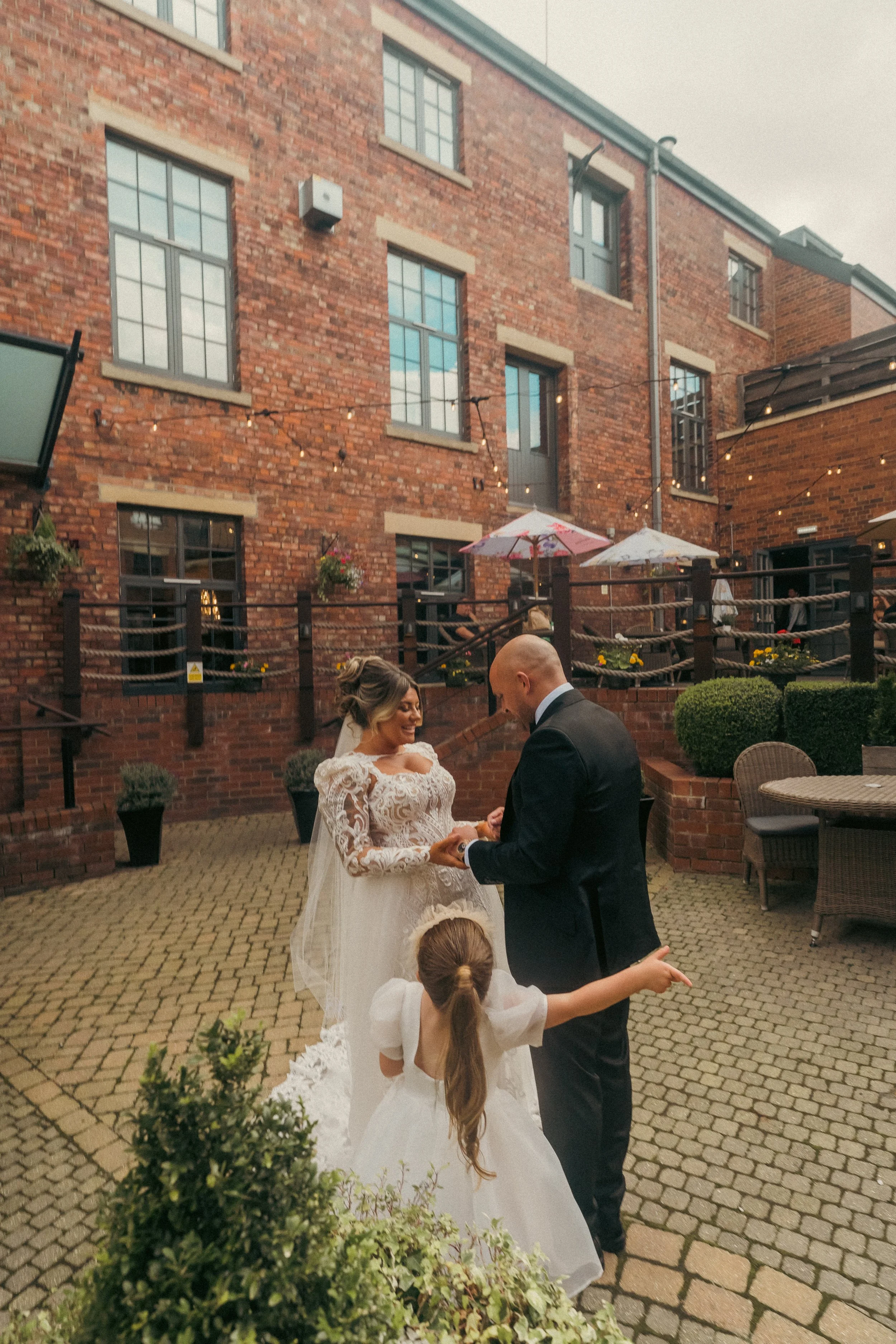 A bride and groom exchange rings during a wedding ceremony outdoors, with a young girl pointing in the foreground, in a brick patio area with string lights and plants.