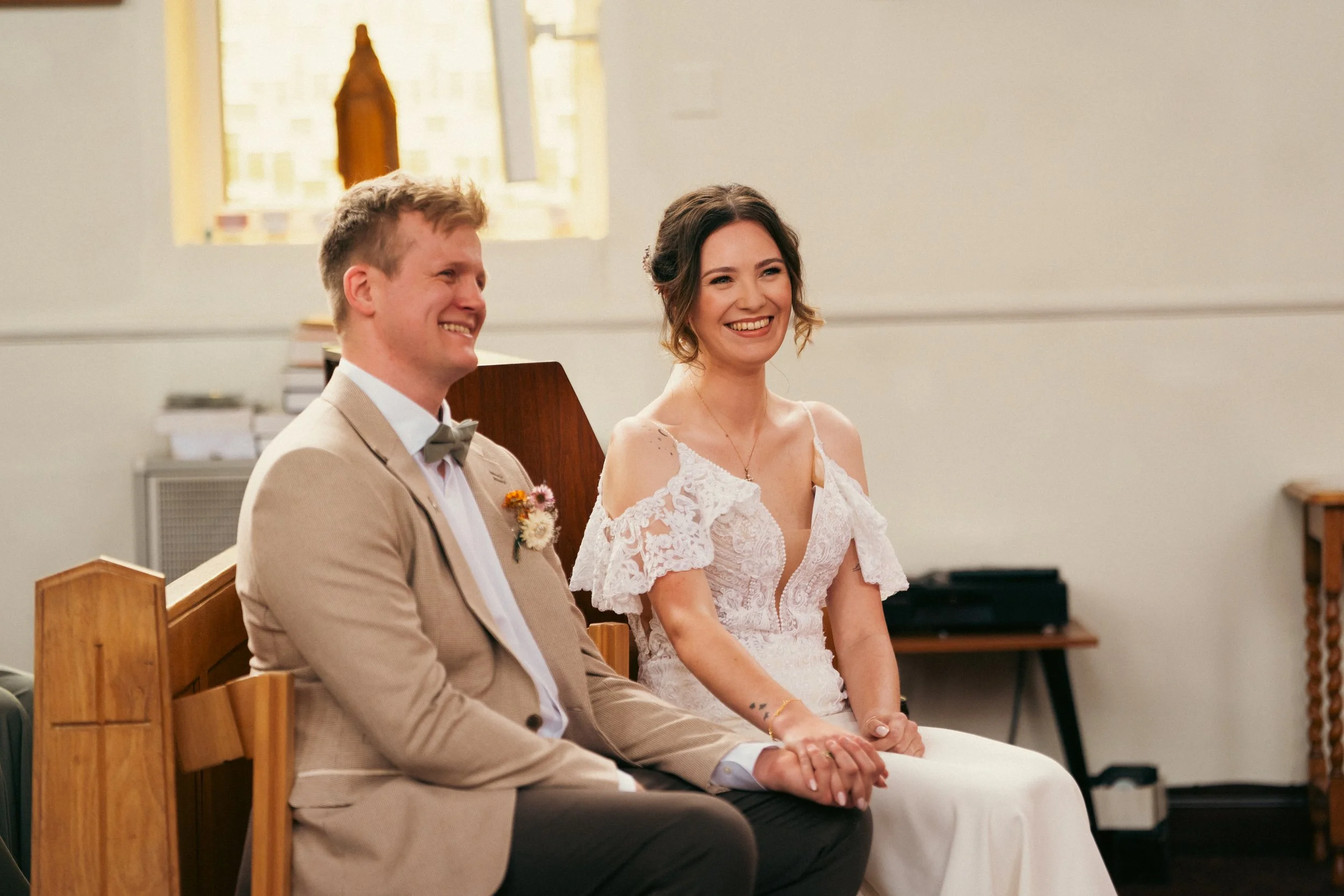 A bride and groom sitting in a church, holding hands and smiling during their wedding ceremony.