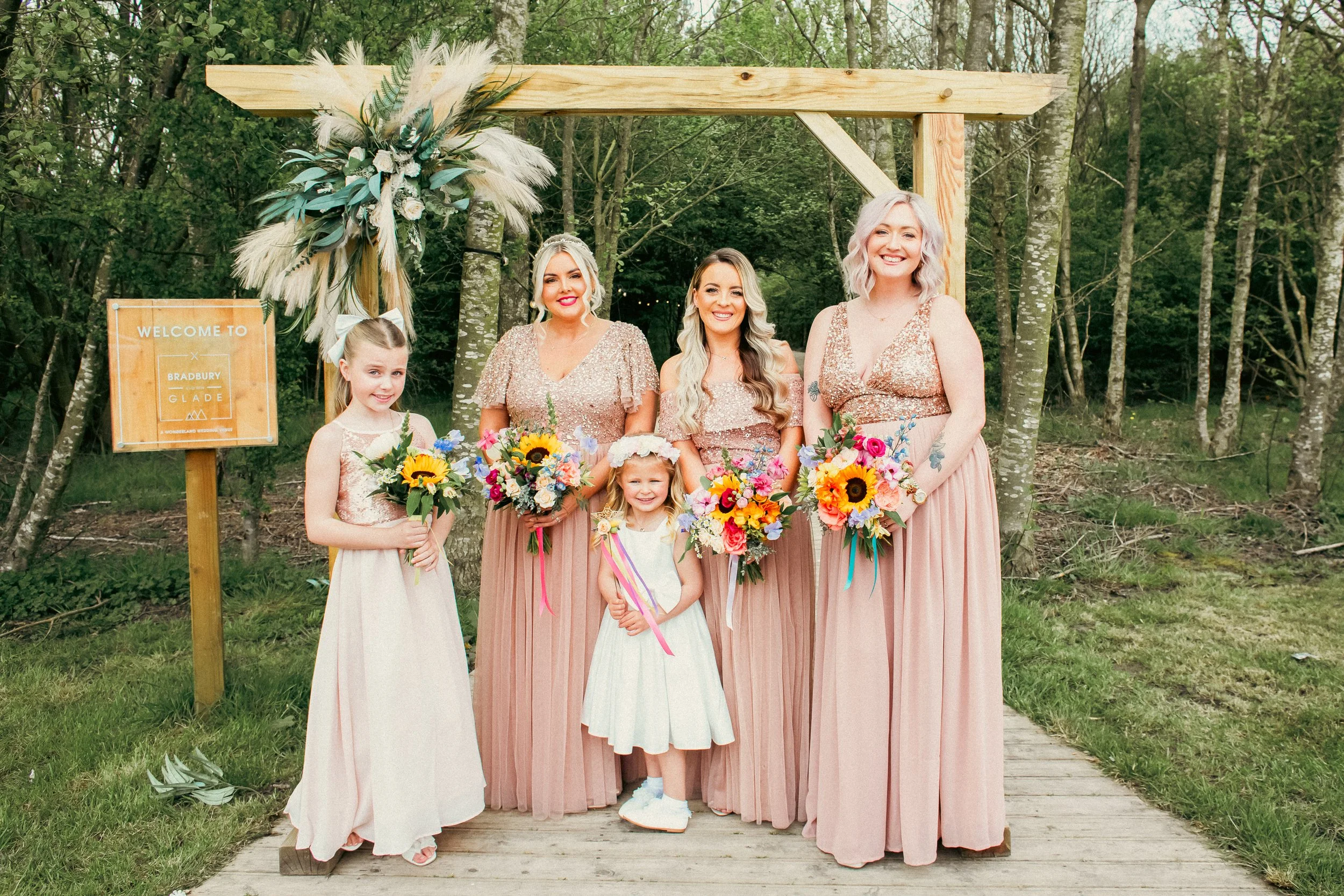 Four women and two young girls standing under a wooden wedding arch outdoors, holding colorful bouquets of flowers, with a sign that reads 'Welcome to Bradbury Glade' in the background.