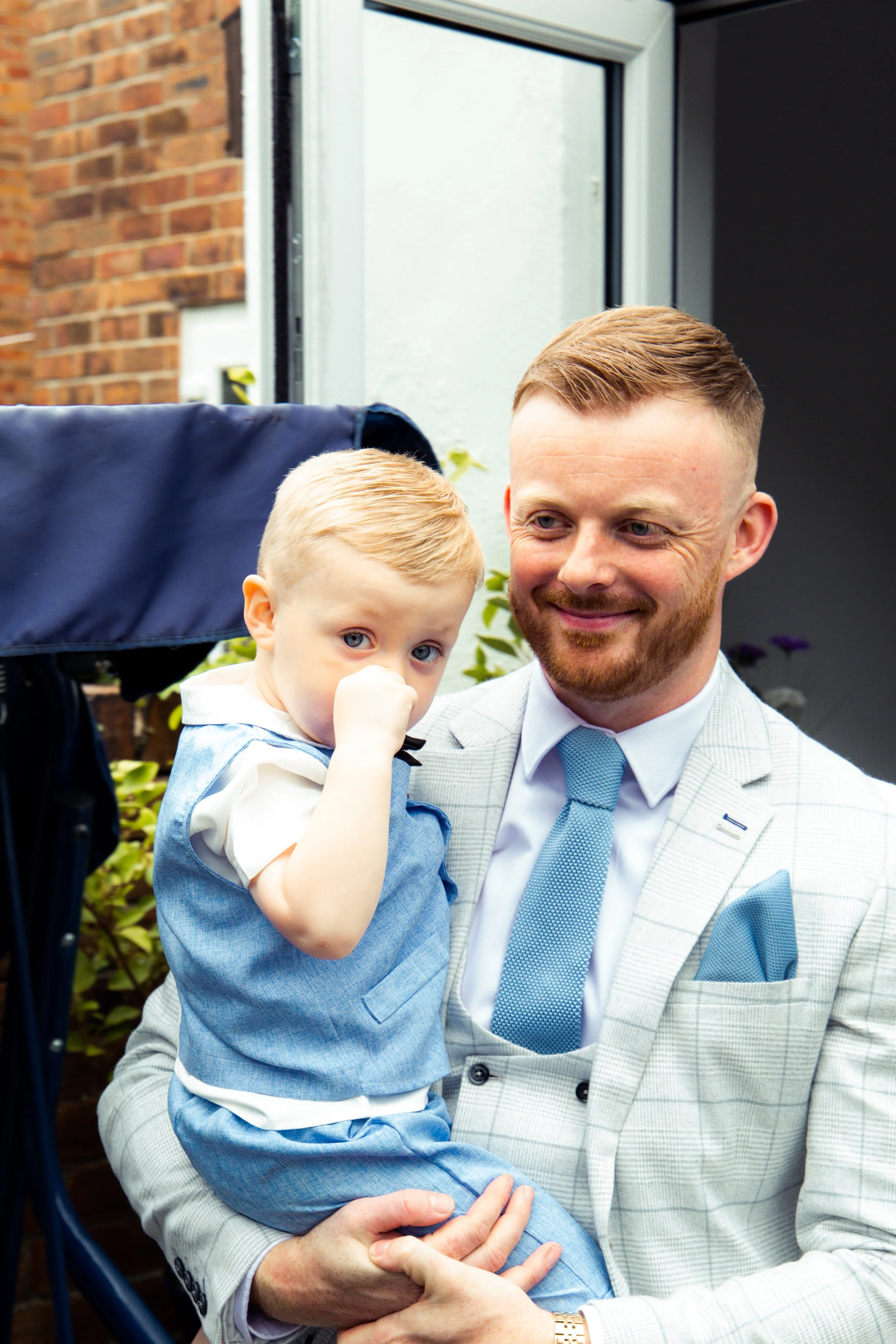 A man in a light-colored suit holding a young boy dressed in blue outside a building.