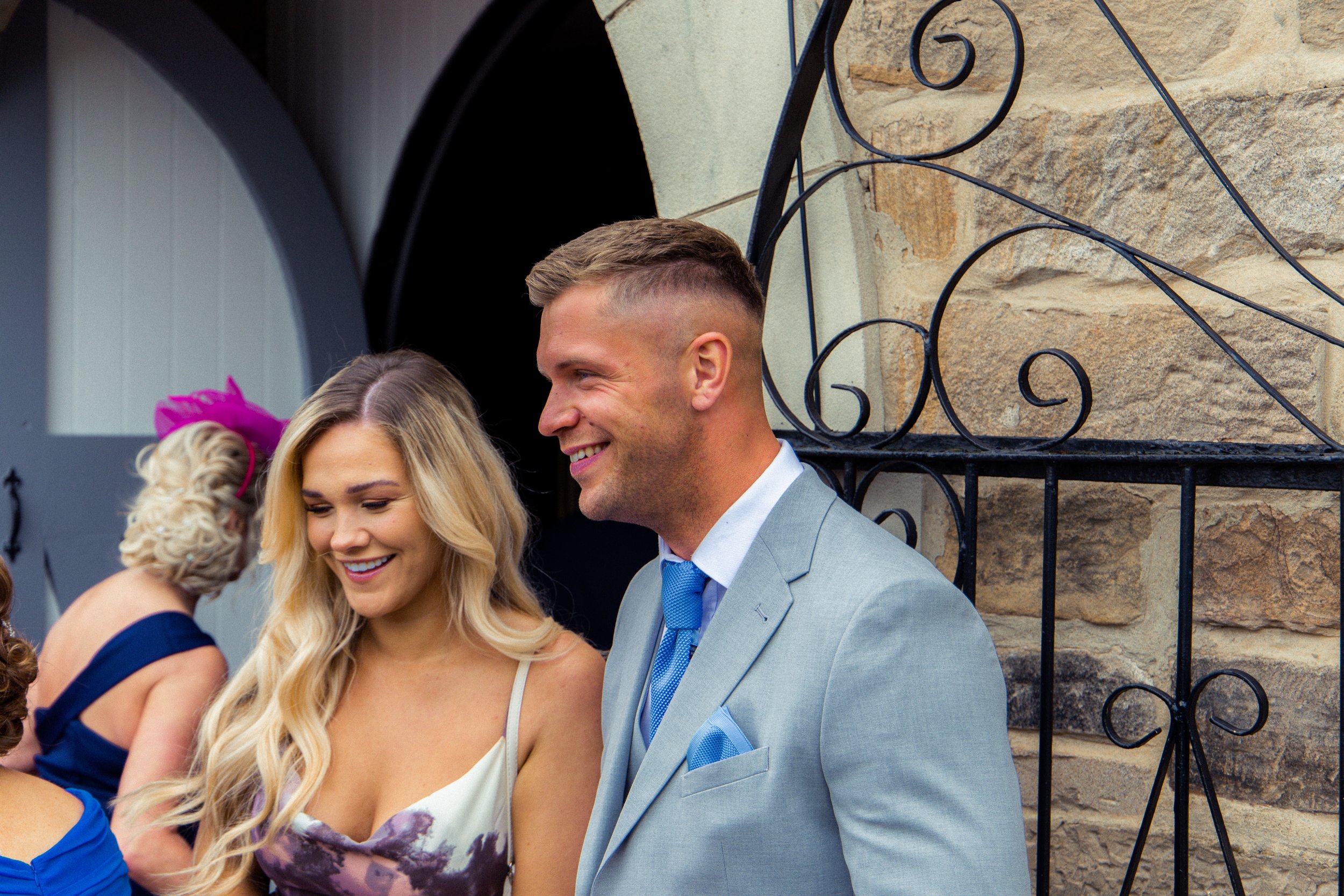 A man and woman are standing outside in formal attire, smiling and looking down, with a stone building and black wrought iron gate behind them.