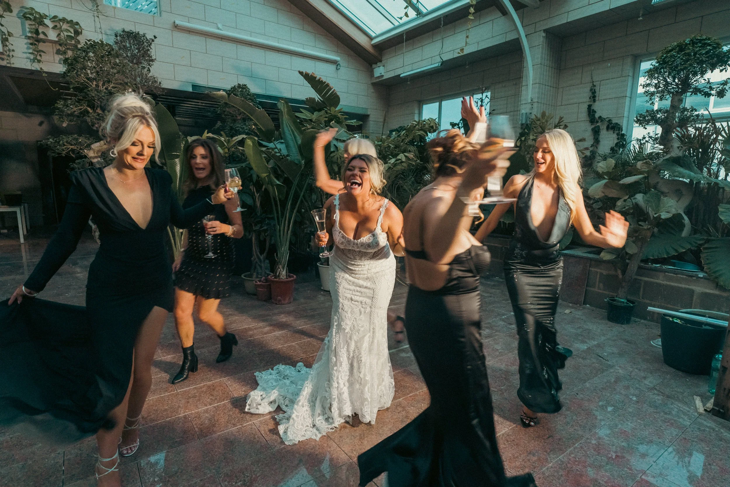 Women dancing and celebrating at a party, with the bride in a wedding dress in the center and several friends holding drinks.