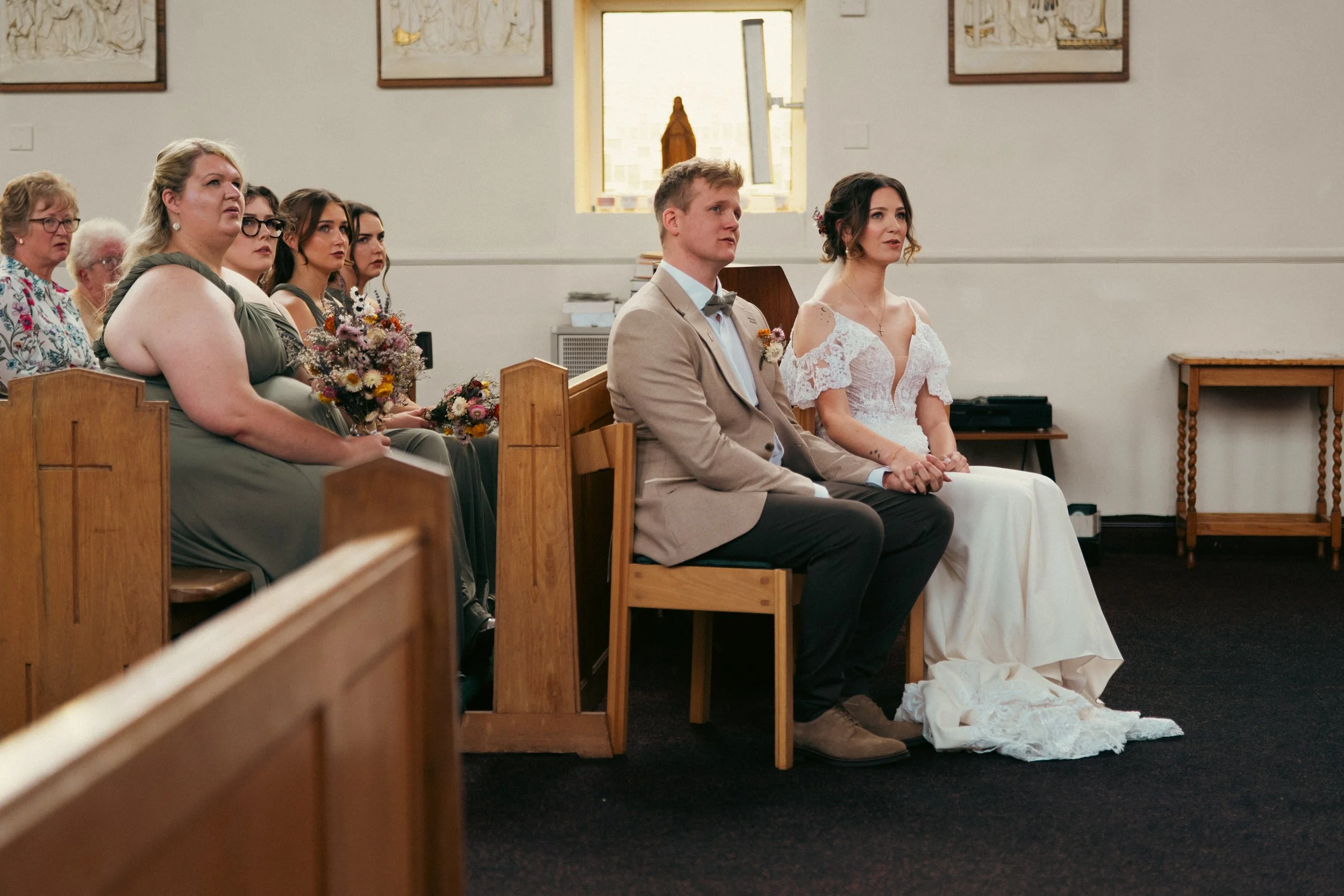 Bride and groom sitting during a wedding ceremony in a church, with bridesmaids and guests seated behind them.