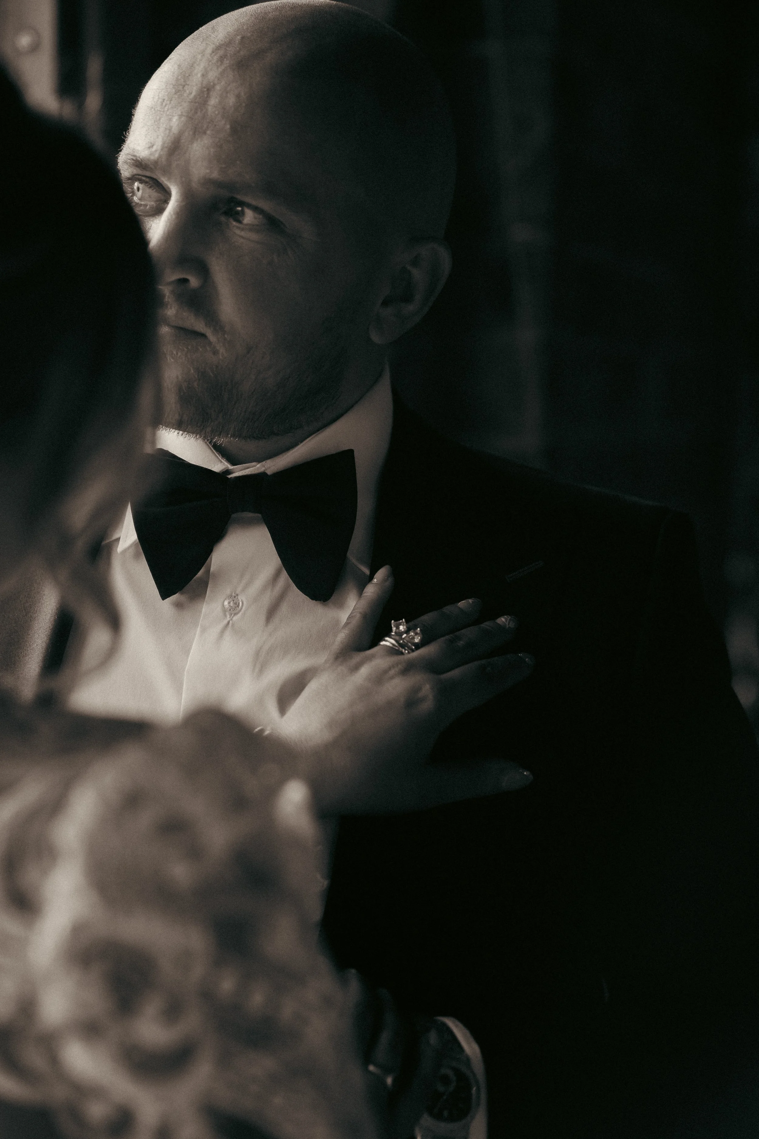 A man in a tuxedo with a bow tie, looking at a woman with her hand on his chest, sharing a tender moment.