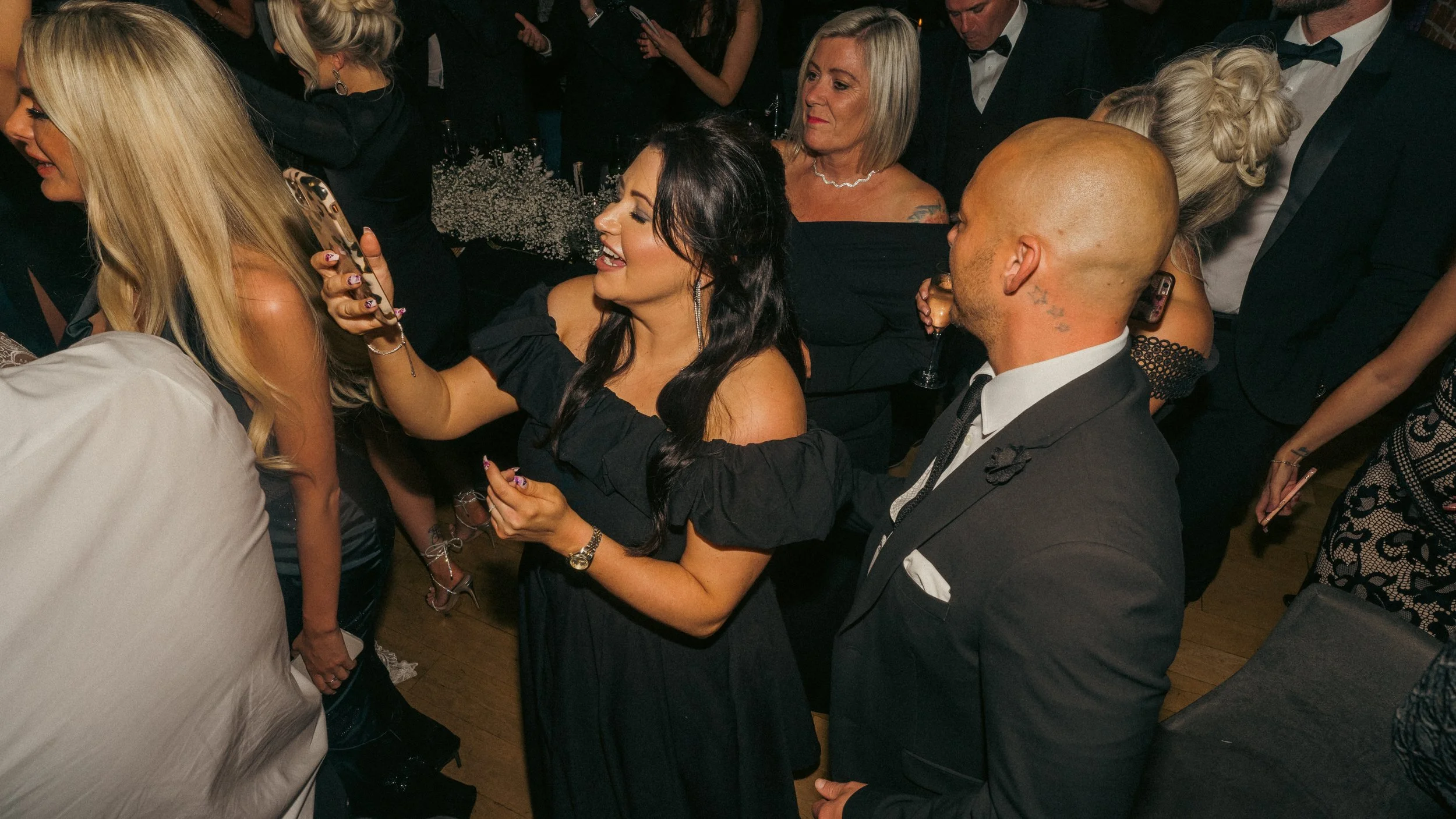People socializing at a formal event, with women in dresses and men in suits and tuxedos, some holding drinks, in a lively atmosphere.