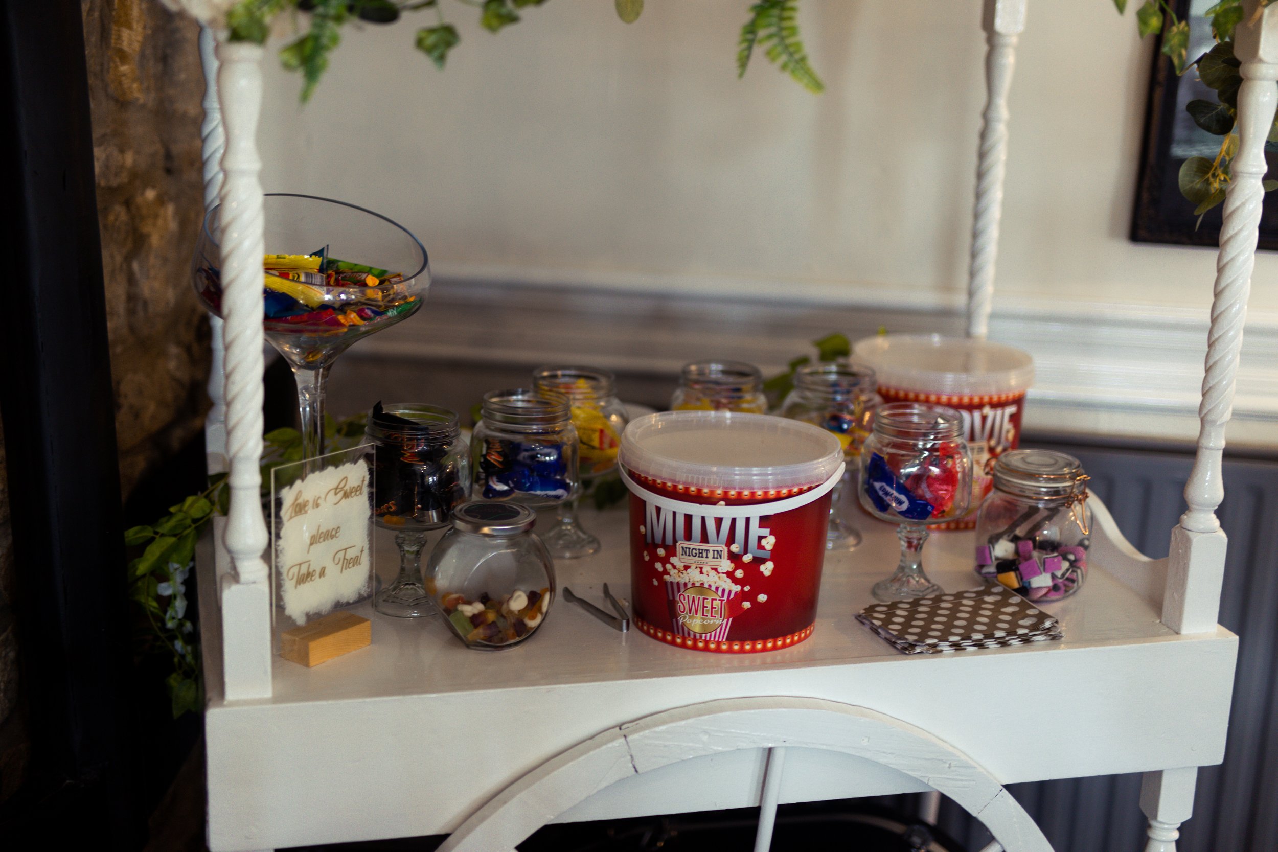 A white table filled with jars of colorful candies and two large buckets labeled "Movie Night in" and "Sweet" for a movie night snack station.